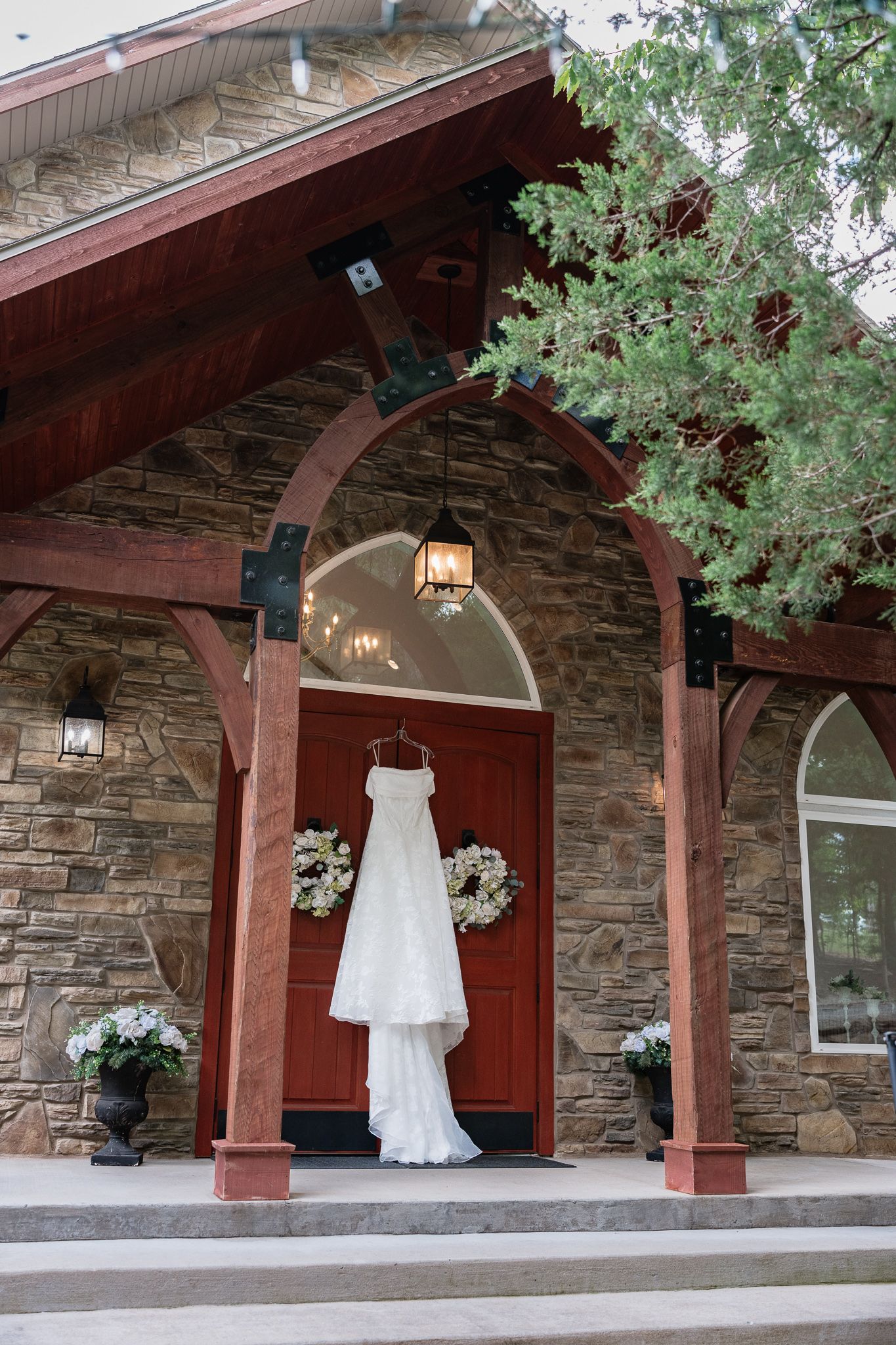 White wedding dress hanging on red door of stone chapel with floral wreaths and warm lantern light.