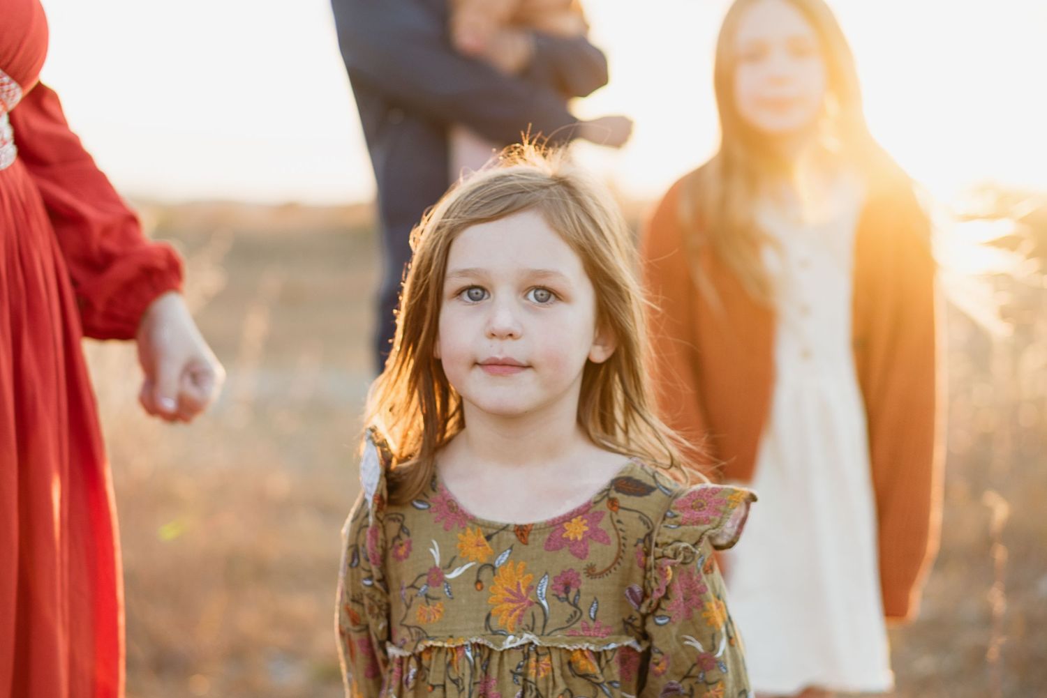 Young girl in floral dress standing in golden field during sunset family photo.