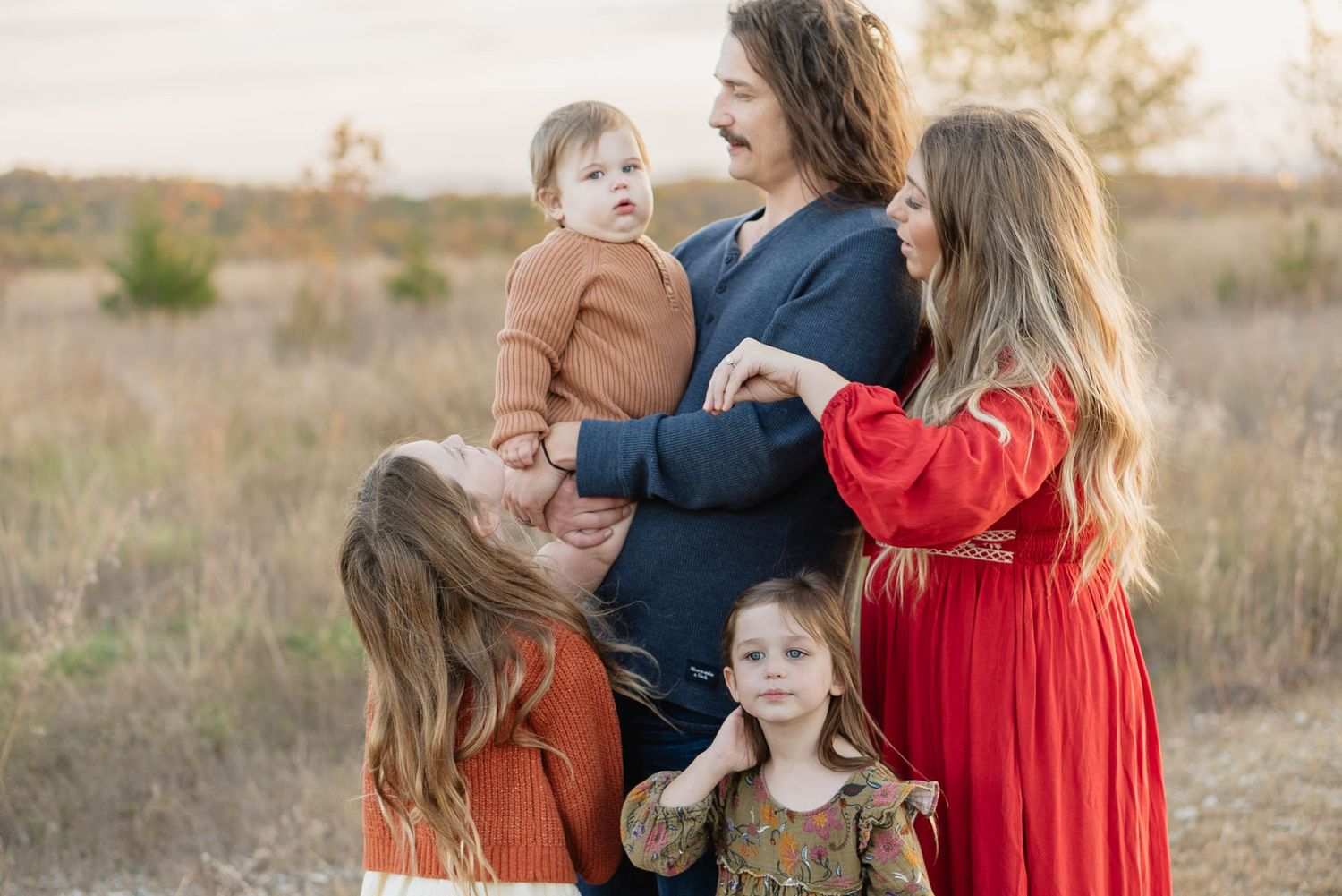 Family portrait with parents and three young children embracing in golden field during autumn outdoor photo session.