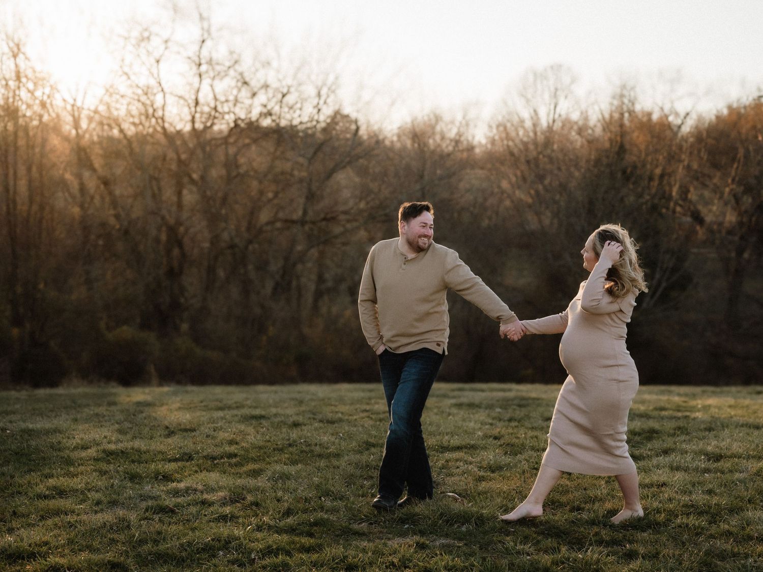 A couple dances hand-in-hand in a sunlit field at golden hour, surrounded by autumn trees.