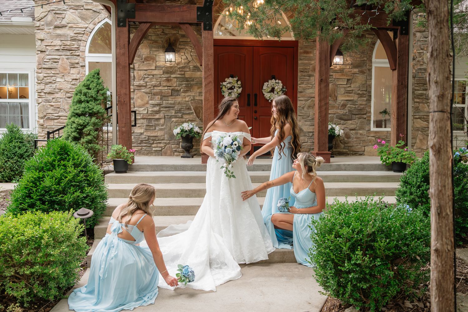 Bride in white gown with bridesmaids in light blue dresses arranging her train outside a stone venue.