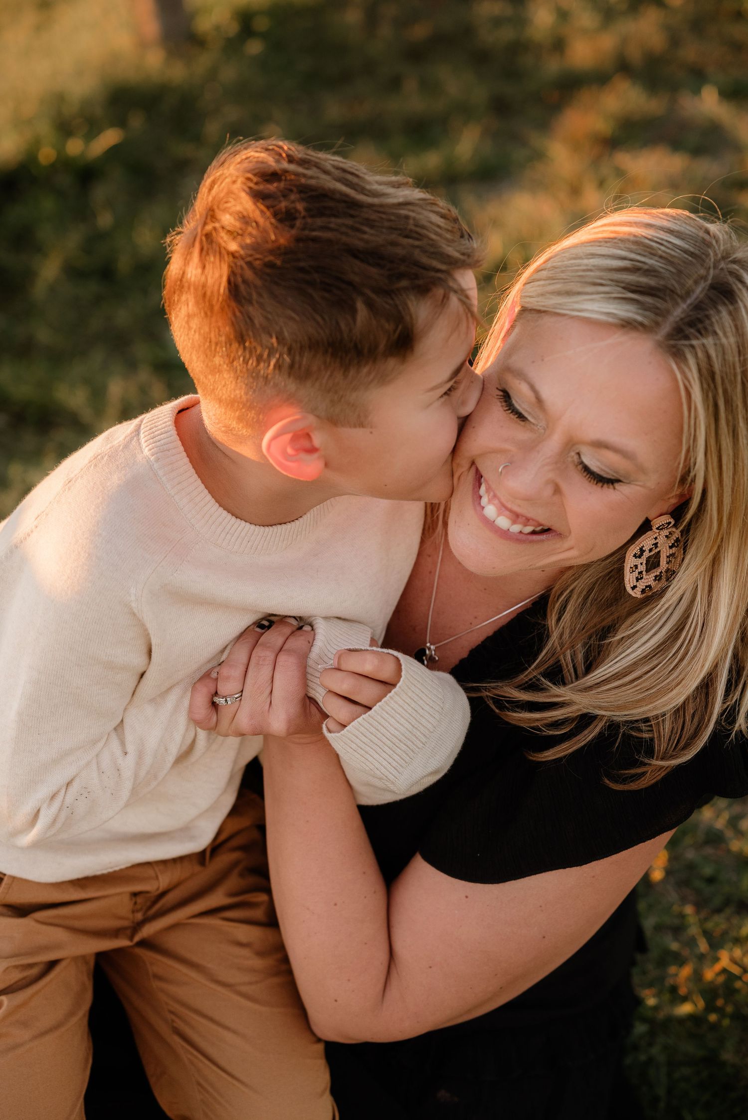 Young boy giving his smiling blonde mother a sweet kiss on the cheek outdoors at sunset.