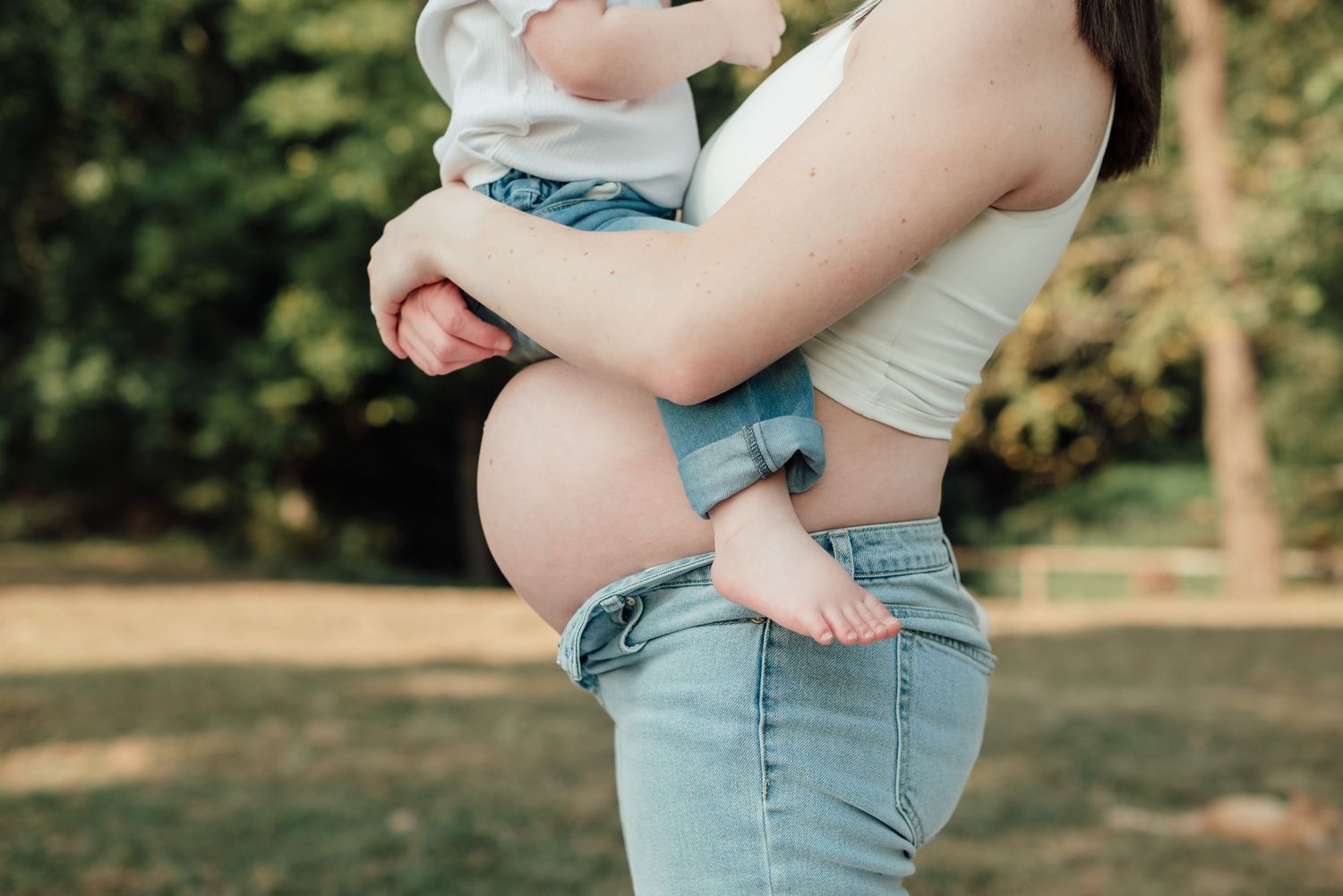 Pregnant woman in white crop top and jeans holding a baby outdoors in a green park setting.