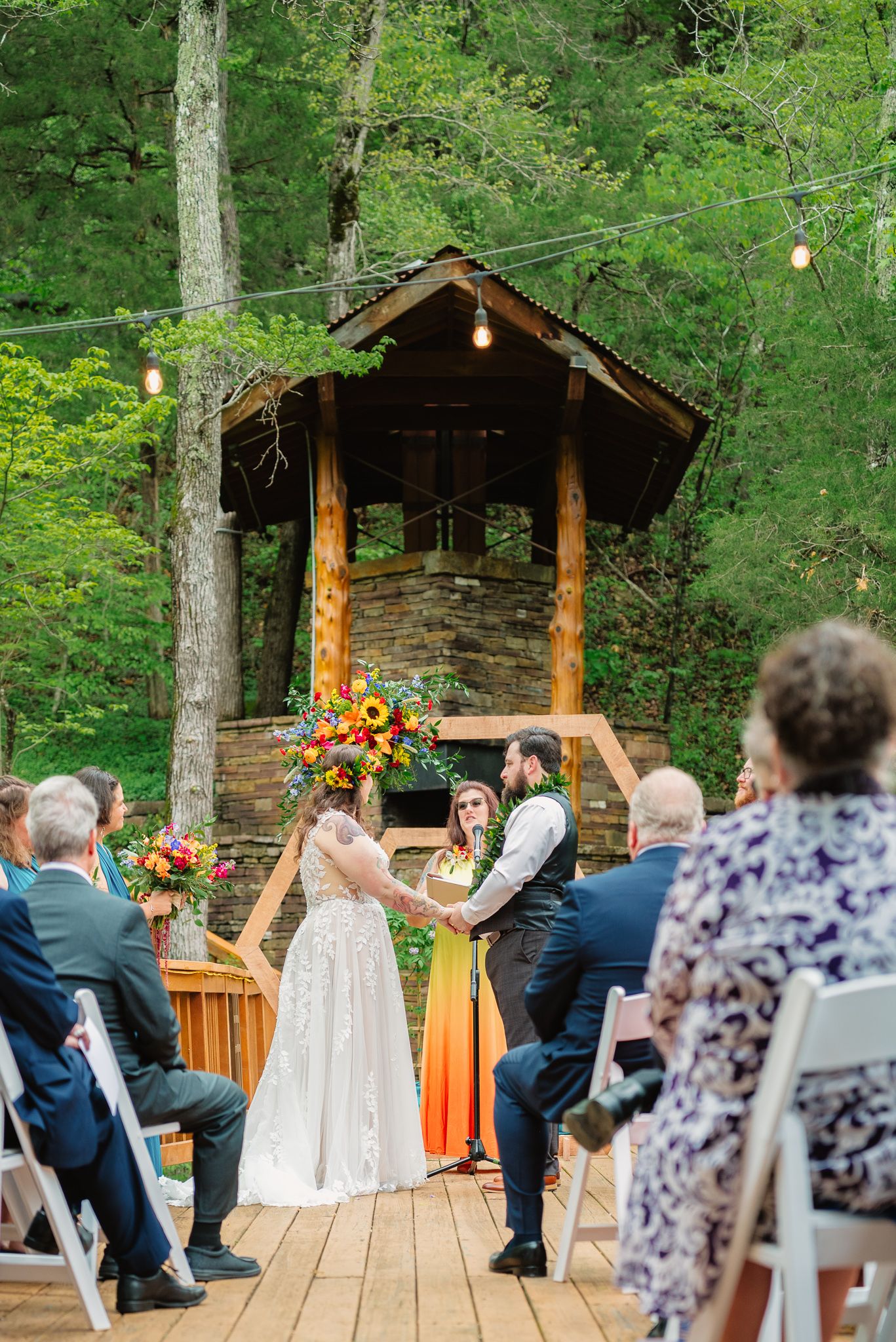Outdoor woodland wedding ceremony on a deck with couple holding hands before an arch, stone fireplace, and string lights.