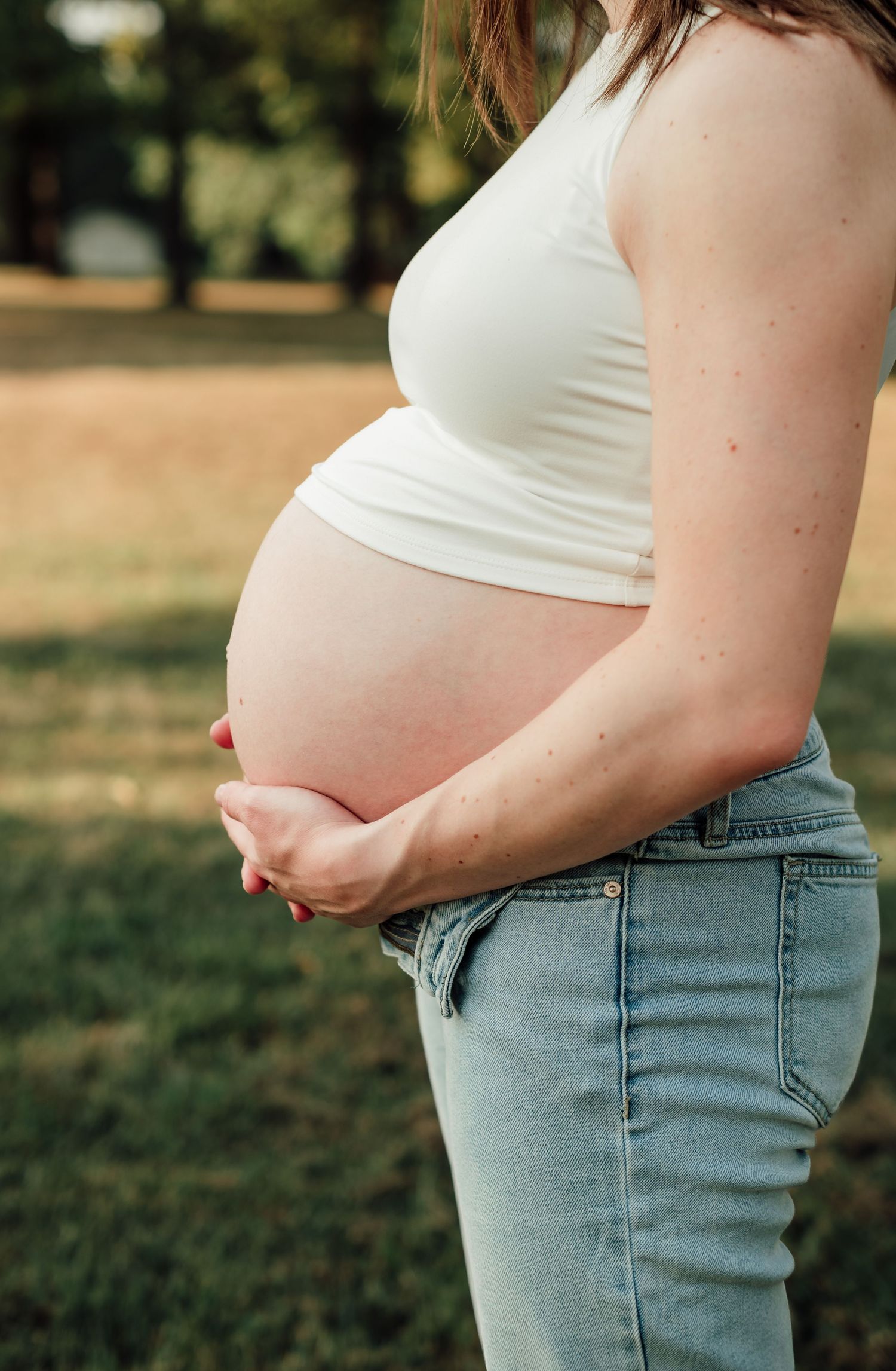 Pregnant woman in white crop top and jeans cradling baby bump outdoors in a park setting.