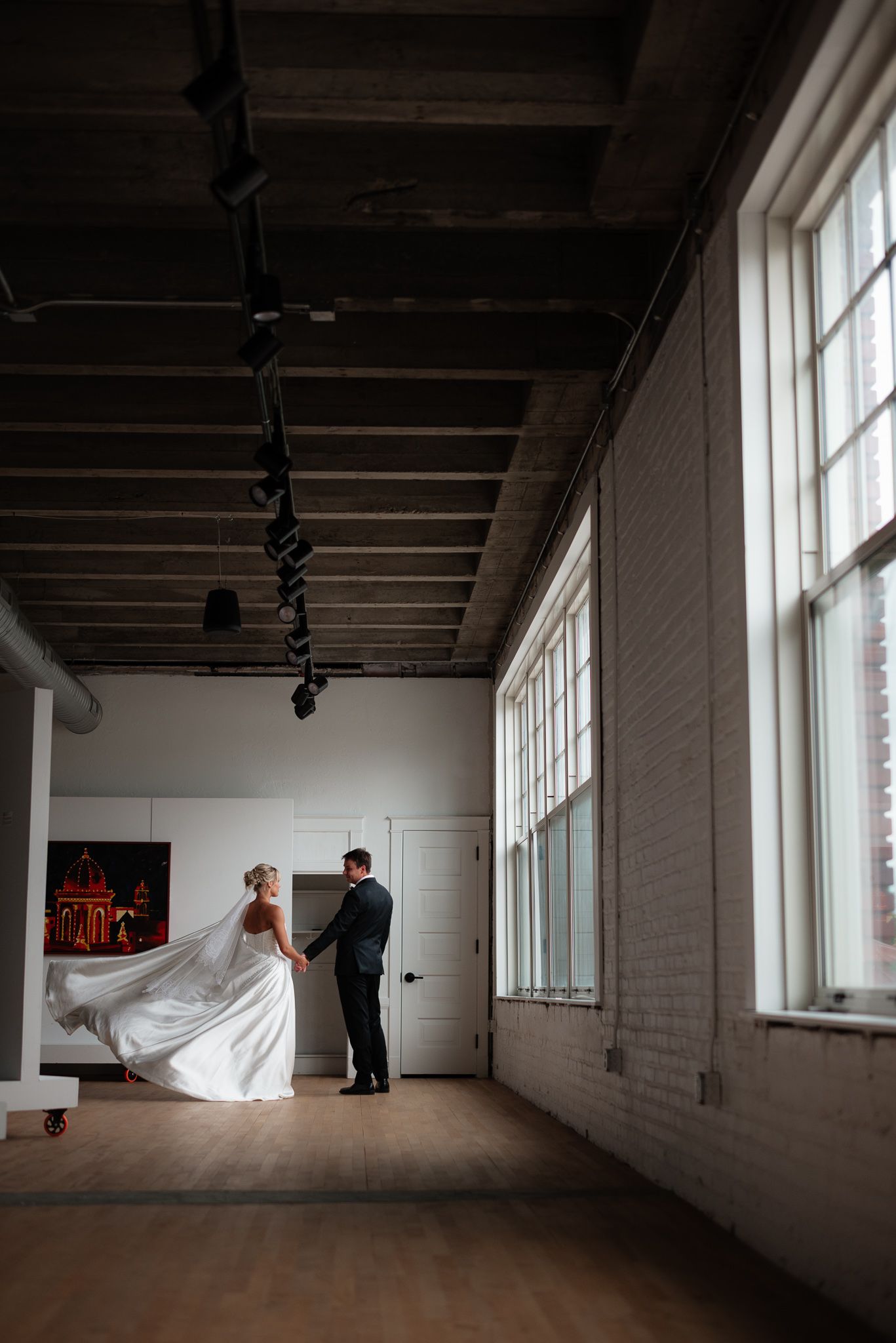 Bride and groom share a romantic first dance in a sunlit industrial wedding venue with exposed brick walls.