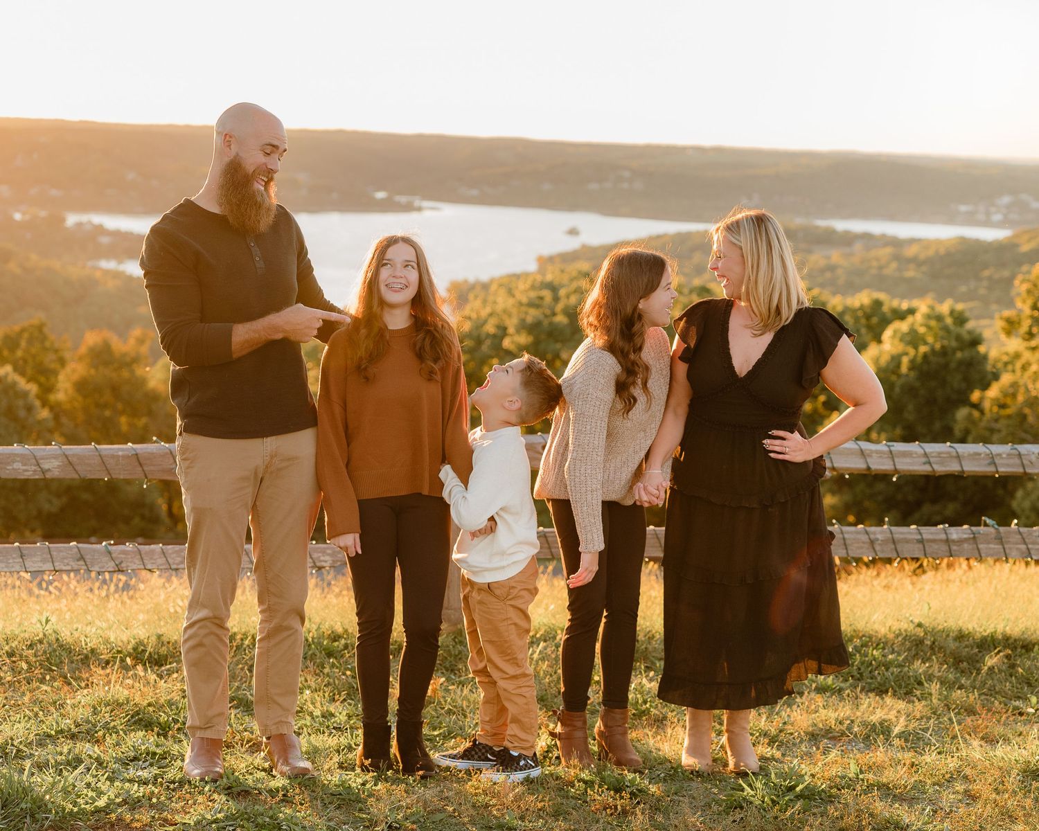 Family of five posing outdoors at golden hour with scenic lake and autumn hills in background.