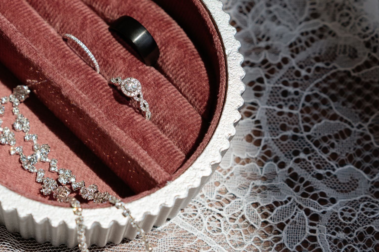 A white ceramic jewelry box with velvet interior holds diamond rings, a black band, and a sparkling necklace on lace fabric.