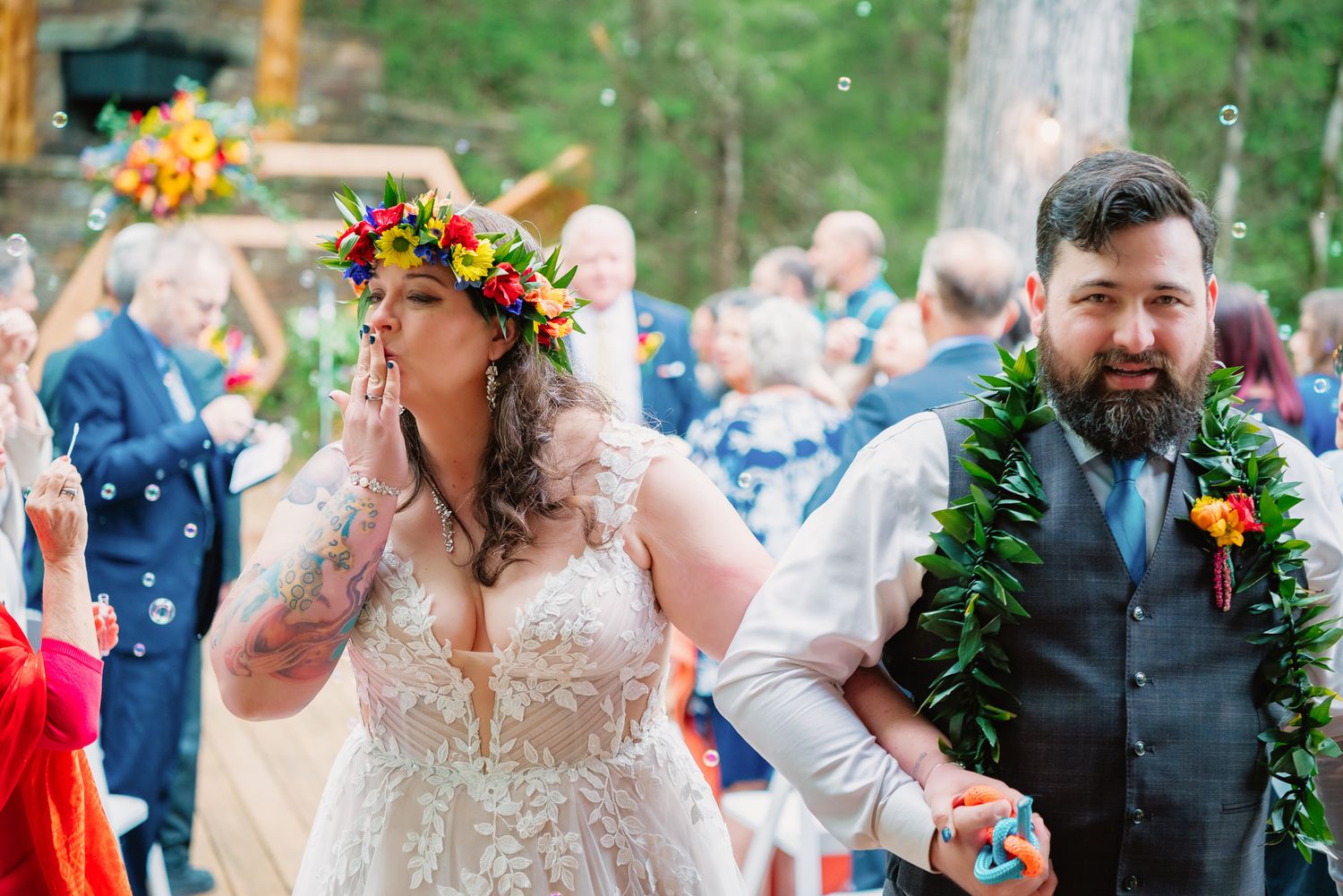 Bride in floral crown blowing bubbles with tattooed groom wearing lei during outdoor wedding reception.
