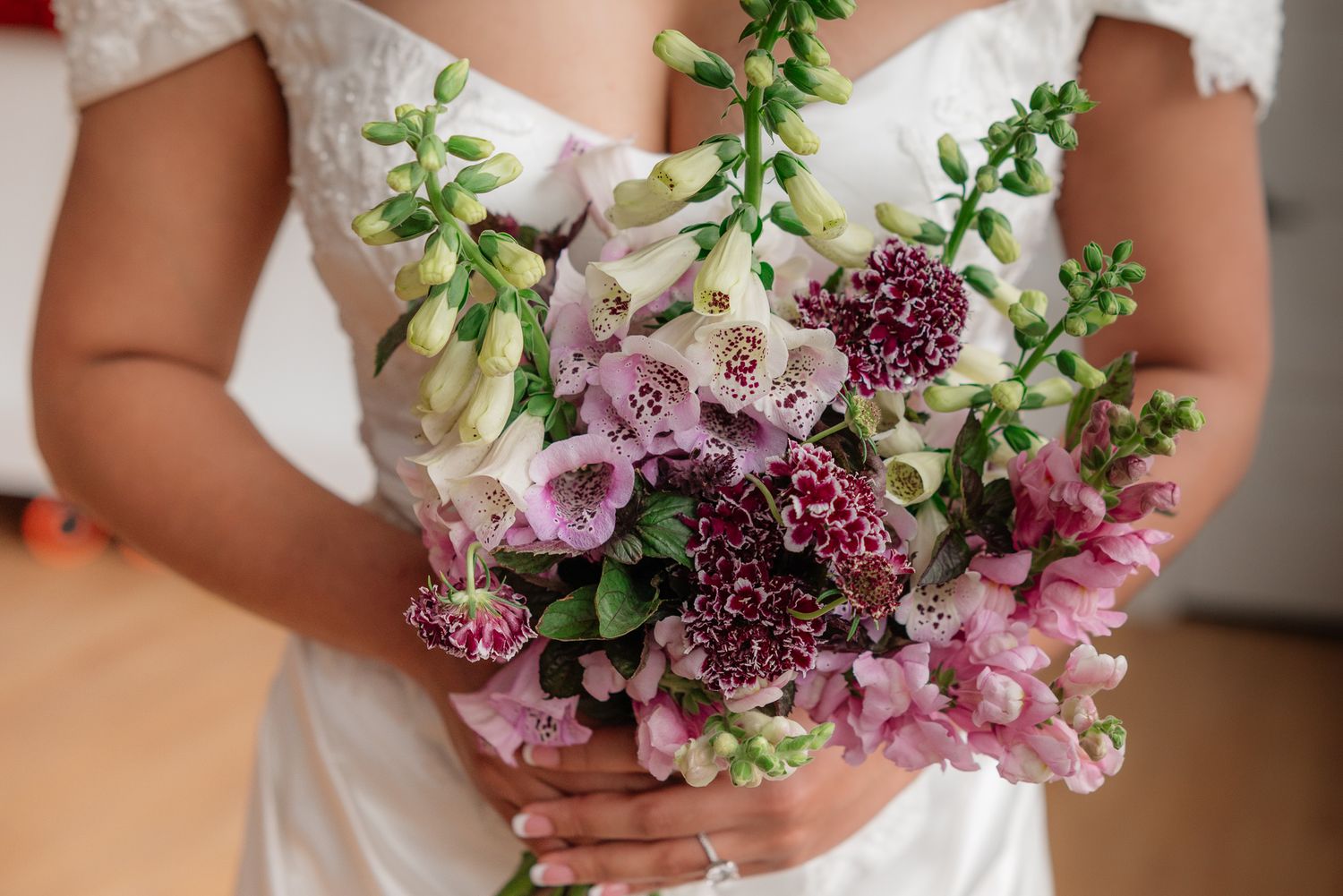 Bride holding a lush wedding bouquet featuring foxglove, pink carnations, and greenery in soft purple and white tones.