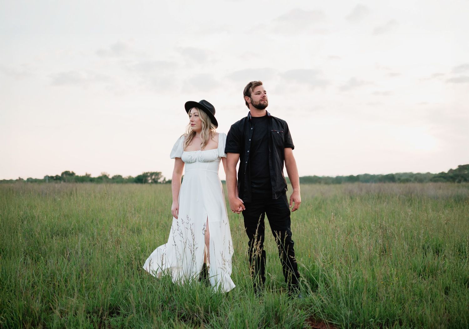 Couple in white dress and black outfit holding hands in a lush green field at dusk.