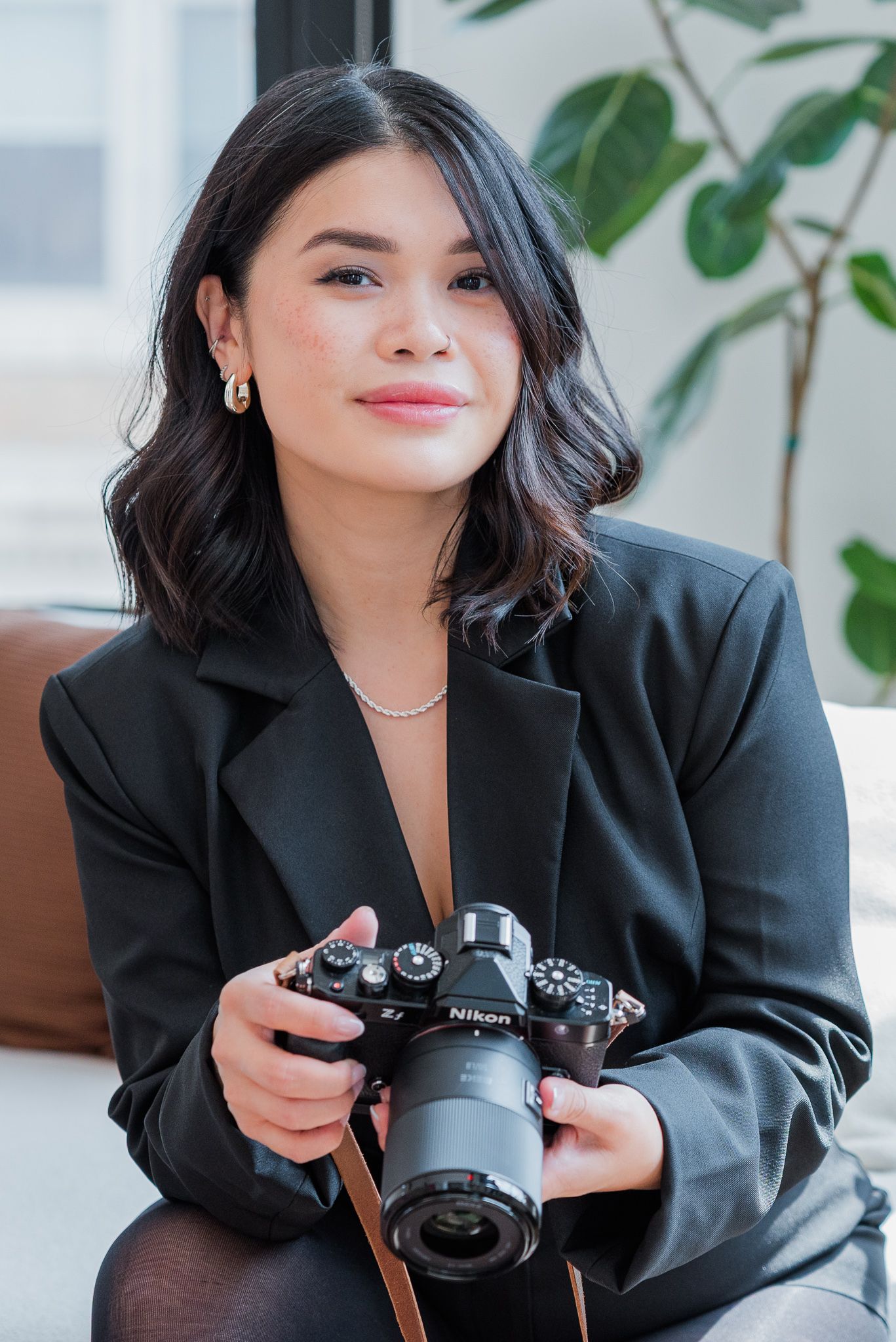 Young woman in black blazer holding a Nikon camera, smiling confidently in a bright modern interior setting.