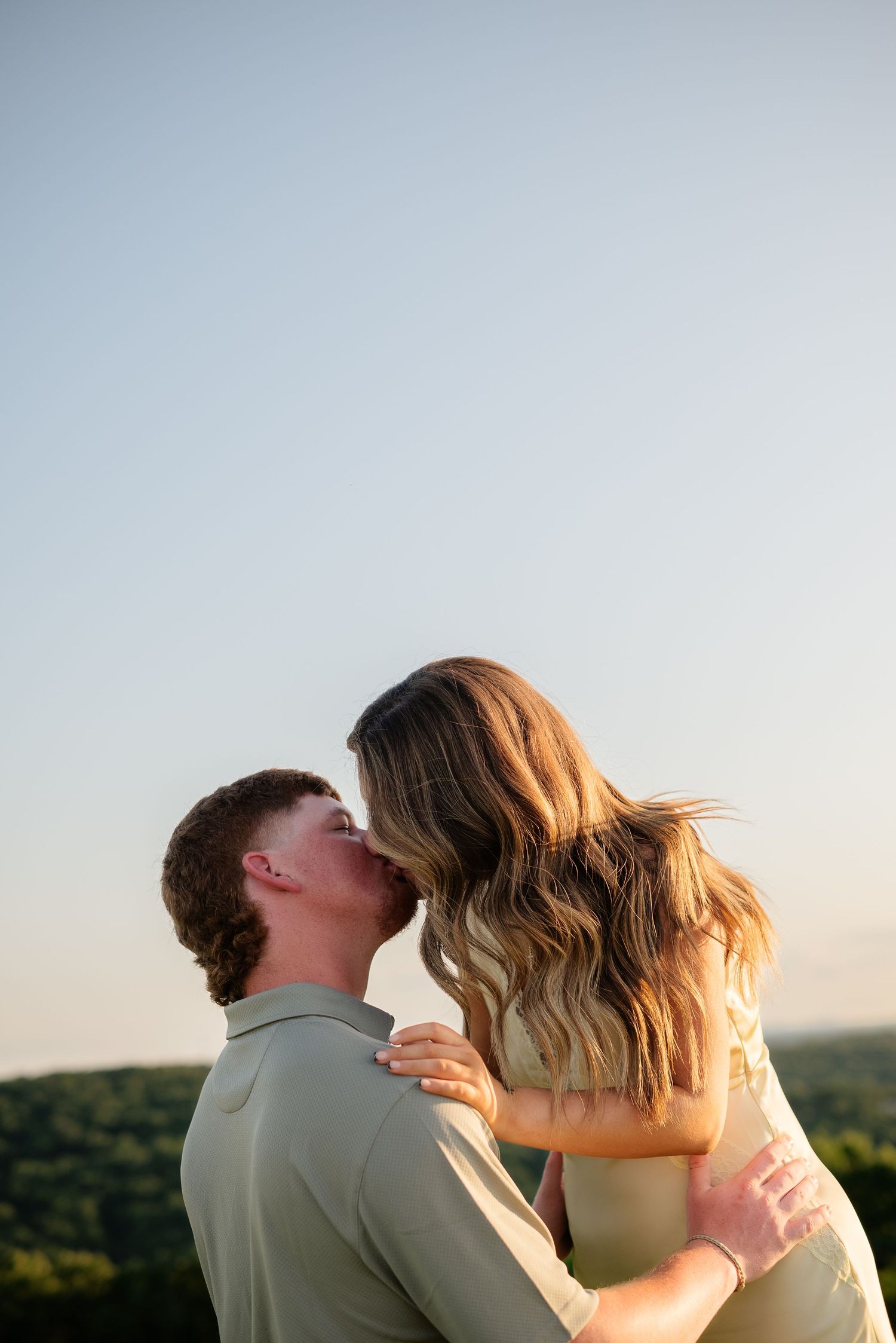A couple shares a romantic kiss outdoors at golden hour with lush green hills and clear sky in the background.