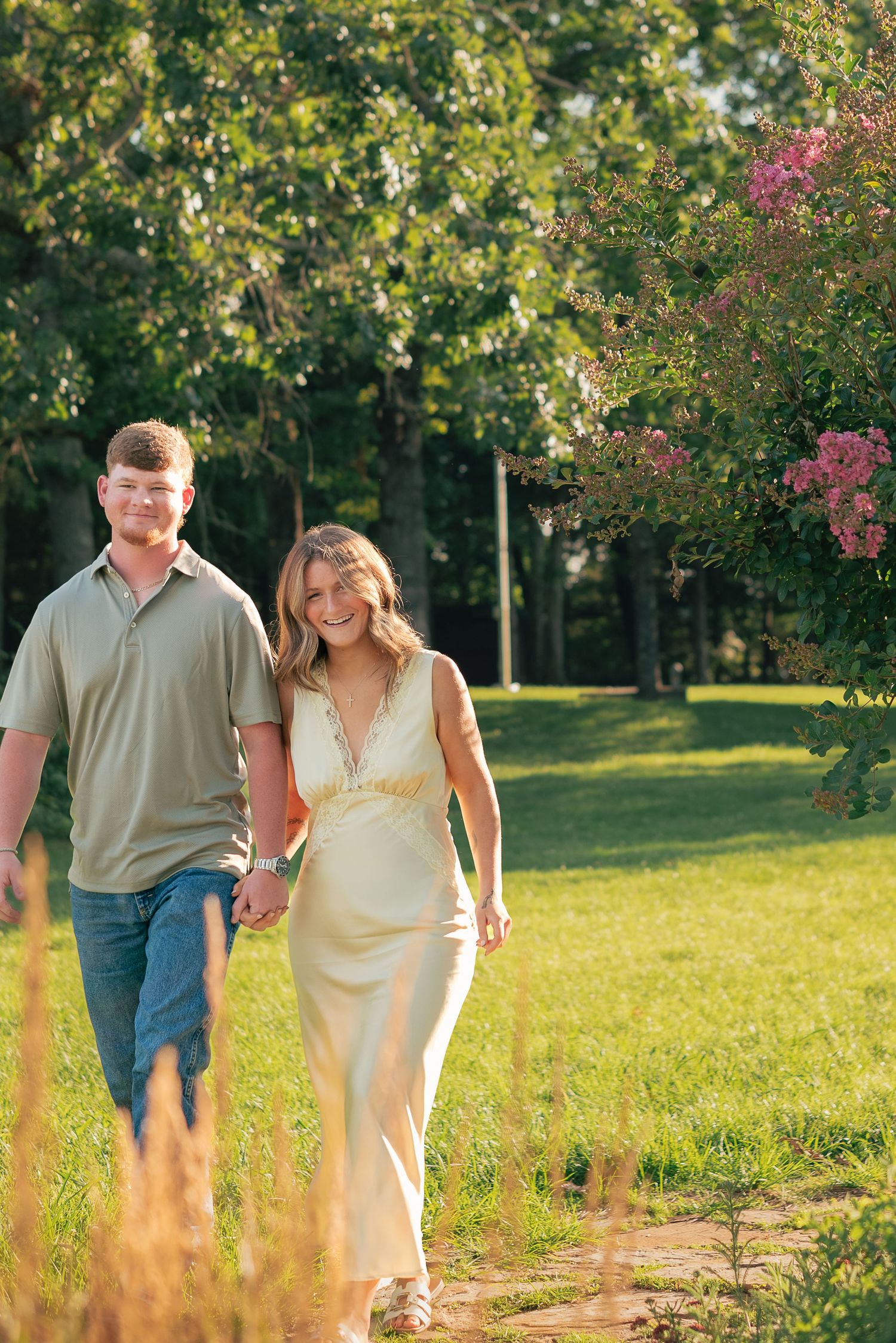 A smiling couple walks hand-in-hand through a lush green park, woman in cream dress, man in olive polo and jeans.