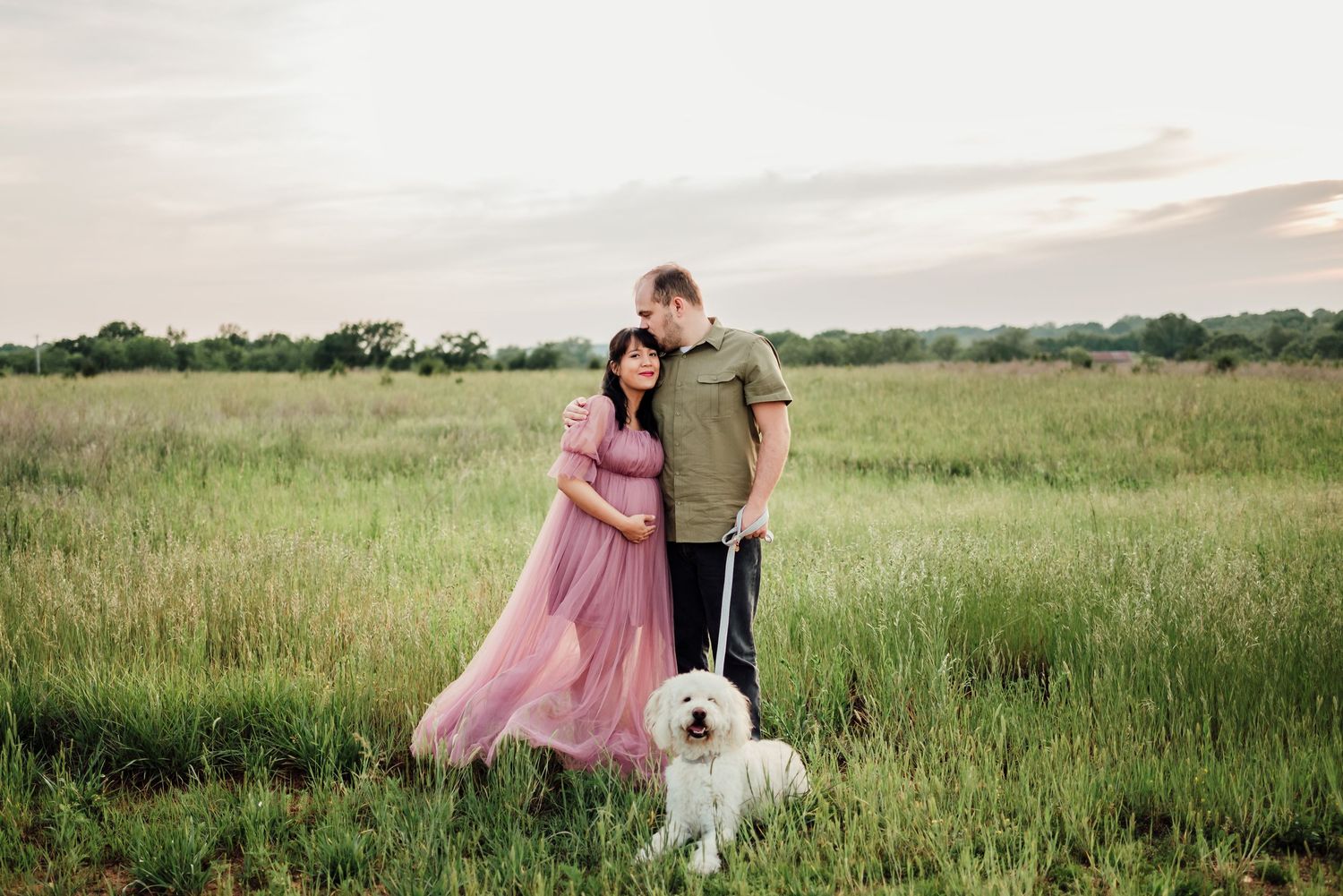 Pregnant couple posing in green field with white fluffy dog, woman in pink maternity gown at sunset.