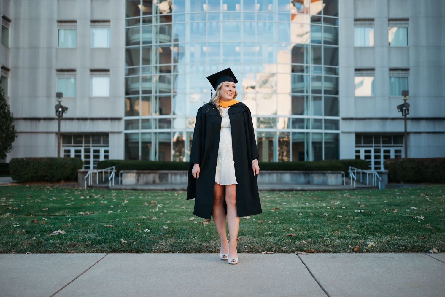 Smiling graduate in black cap and gown poses outdoors in front of a university building on a sunny day.