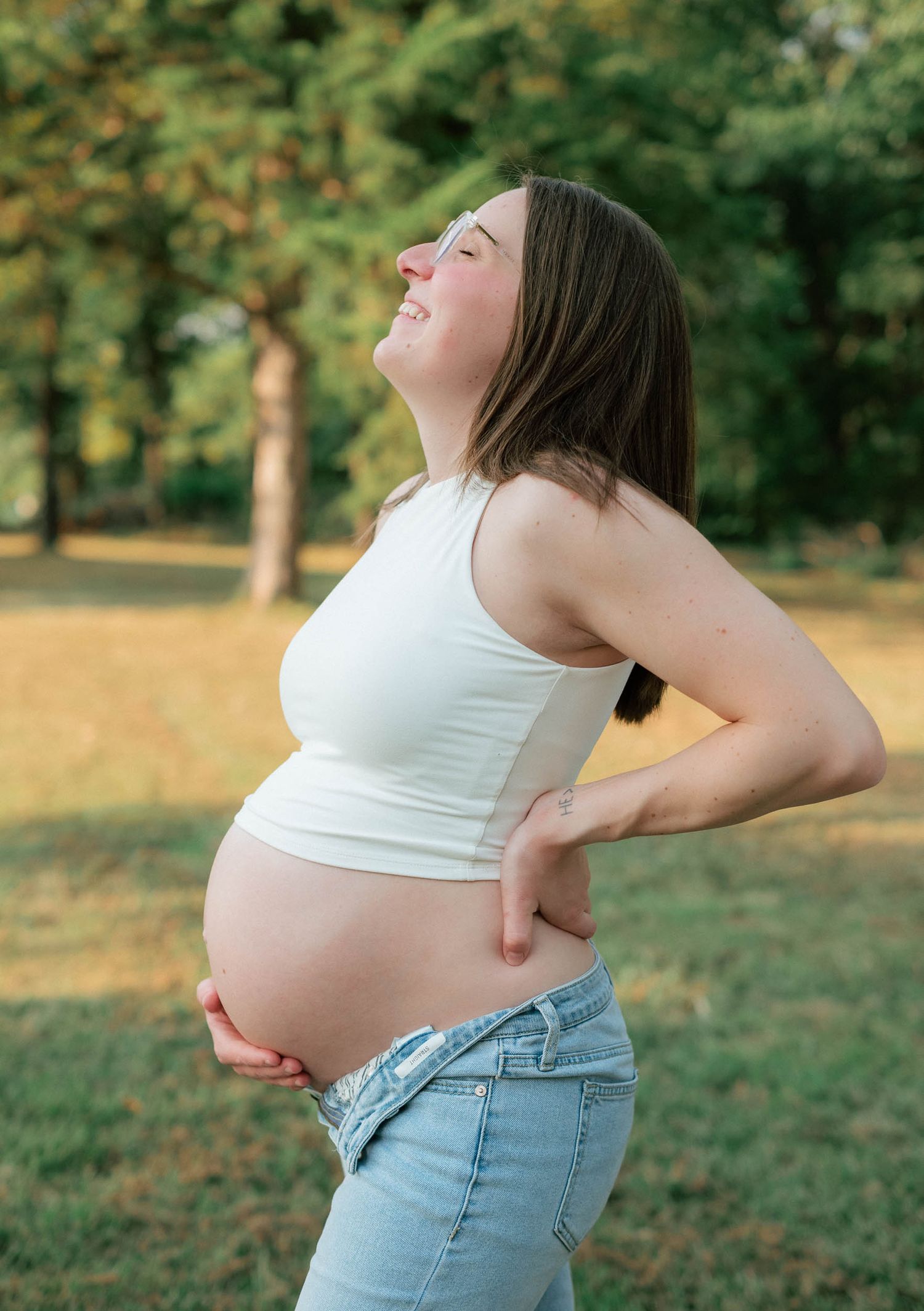 Pregnant woman in white crop top and jeans holding her baby bump outdoors in a sunny park.