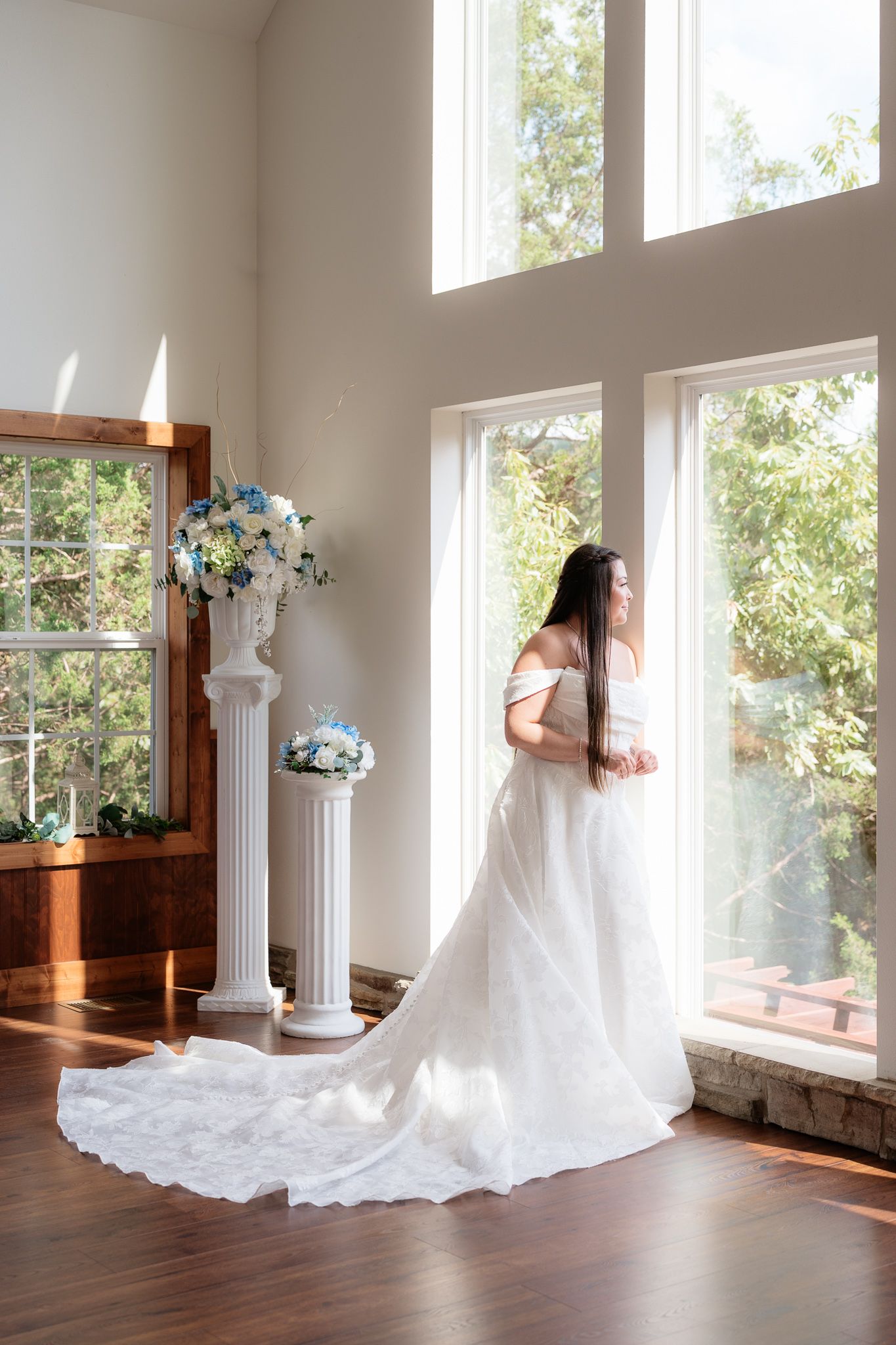 Bride in white off-shoulder gown with long train stands by tall windows, with white floral arrangements on pedestals nearby.