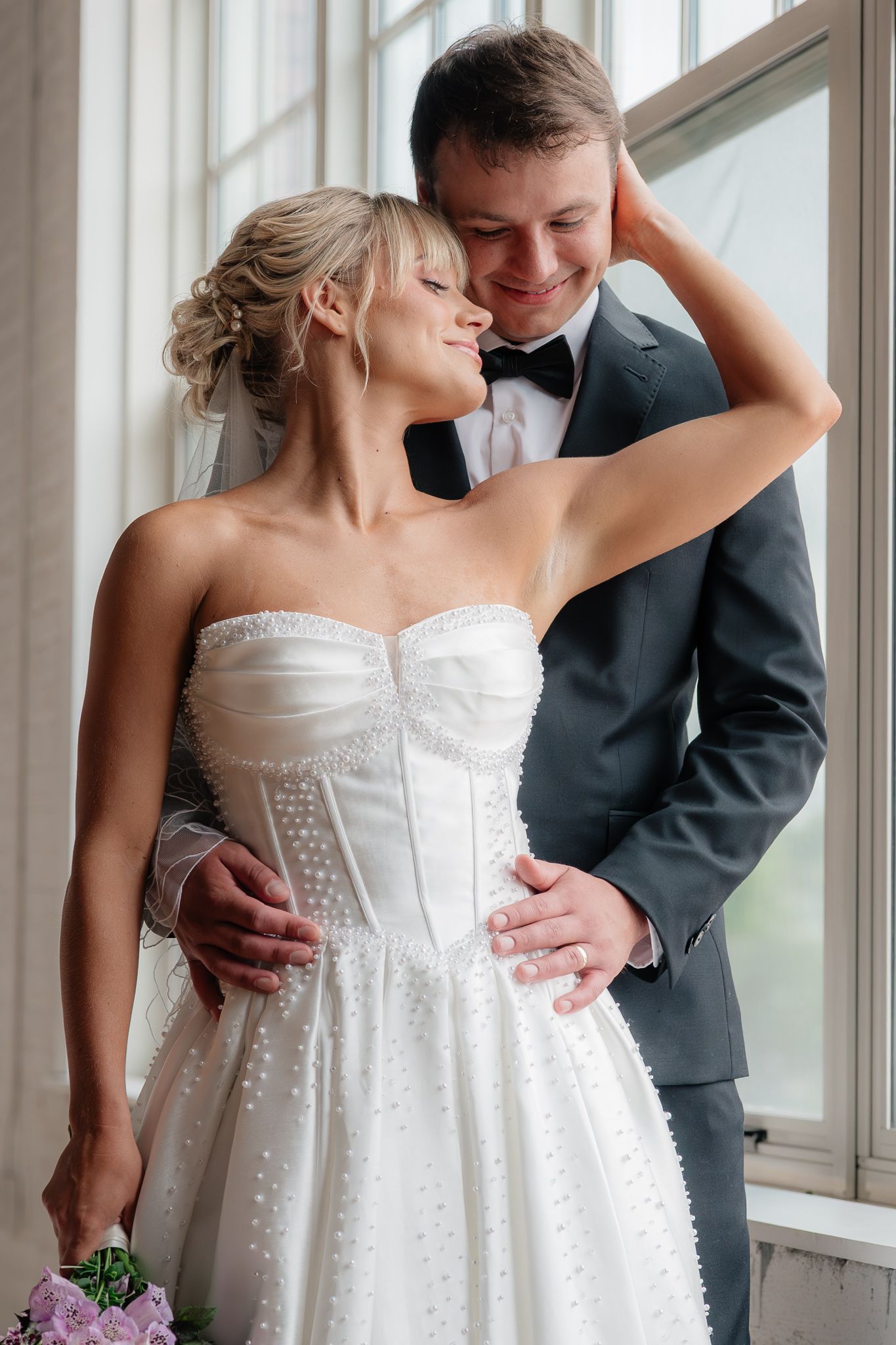 A smiling bride in a strapless white gown holds a bouquet as her groom in a gray suit embraces her by a bright window.