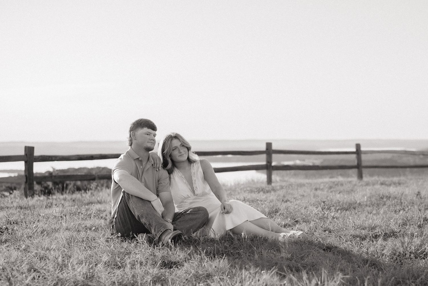 Black and white photo of a couple sitting together on grass near a wooden fence with ocean views.