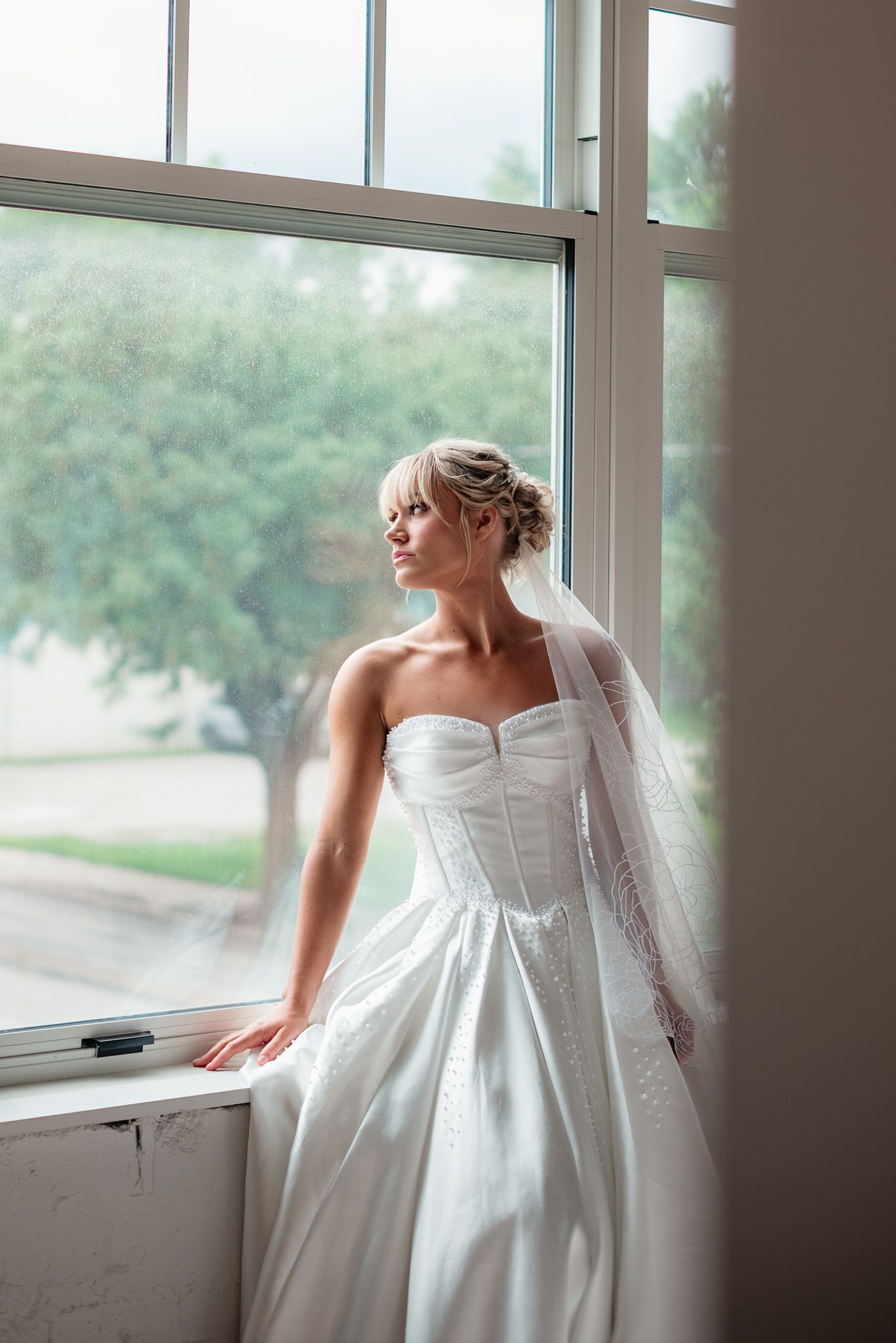 Elegant bride in strapless white gown and veil gazes out large window on her wedding day.