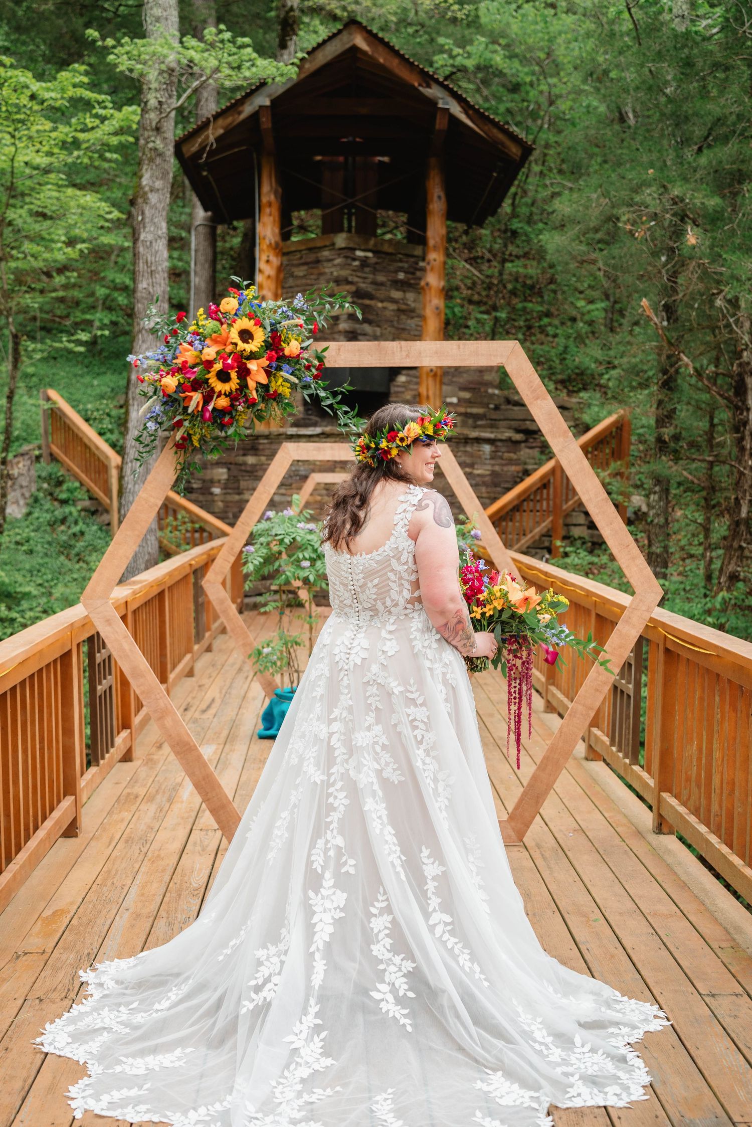 Bride in lace wedding gown with floral crown holds bouquet on wooden bridge before rustic forest chapel.
