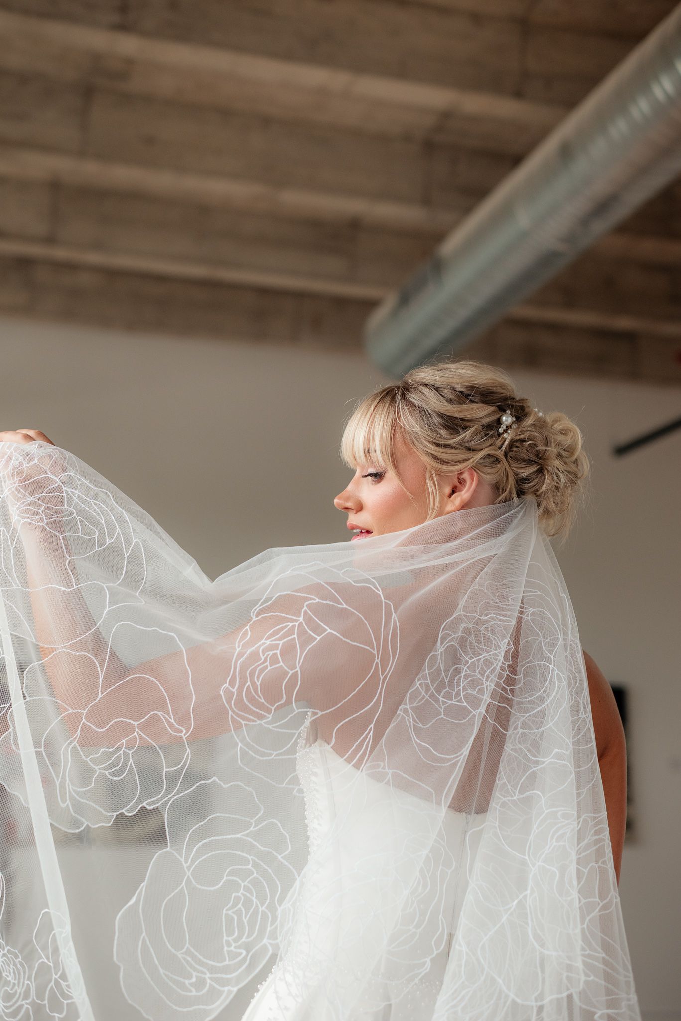 Bride in white wedding dress holding floral lace veil, elegant updo with hair accessory, industrial venue backdrop.