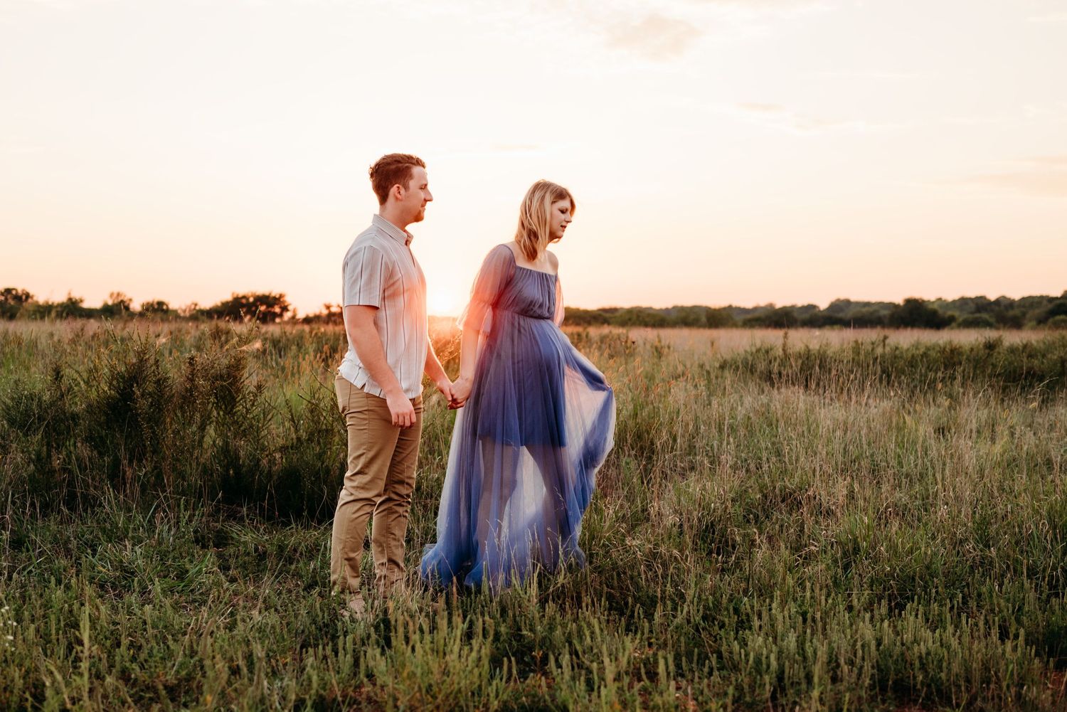Couple holding hands in a golden field at sunset, woman wearing a flowing blue maxi dress.