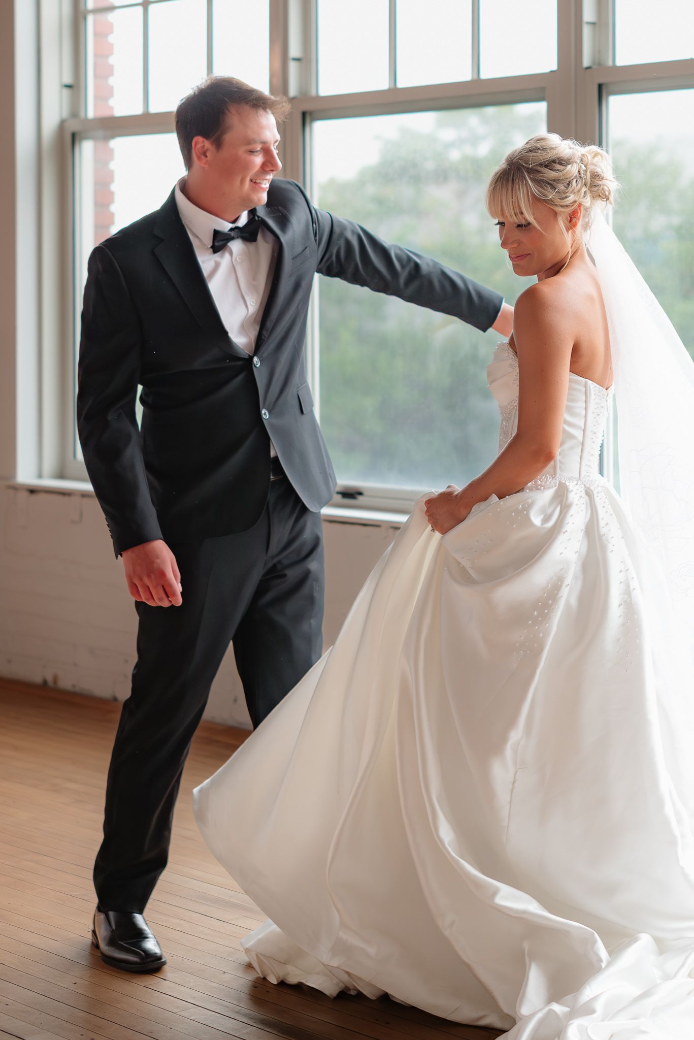 Bride and groom share first dance in elegant white gown and black tuxedo by large windows.
