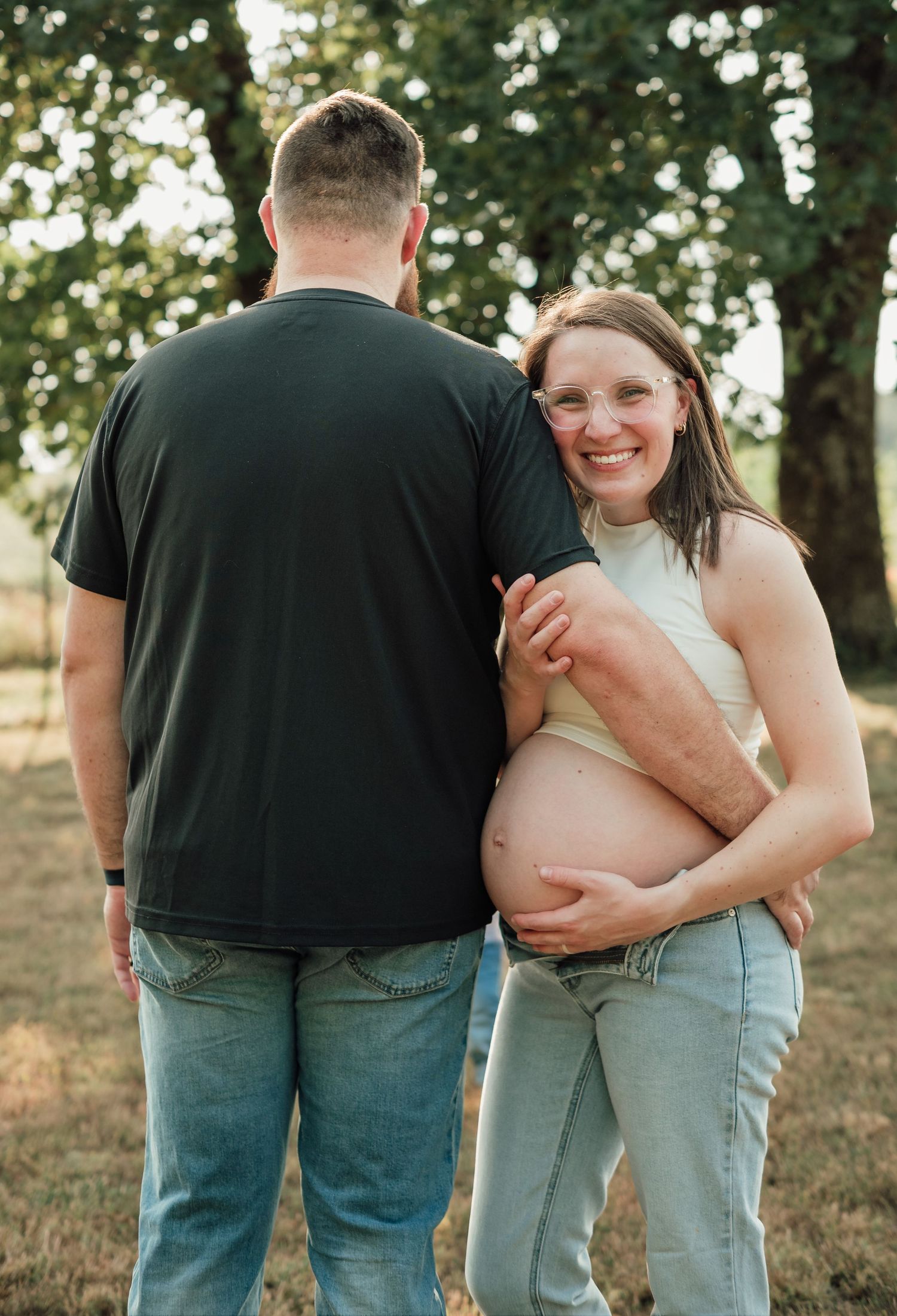 Smiling pregnant woman in jeans cradles baby bump beside man in black shirt during outdoor maternity photo session.