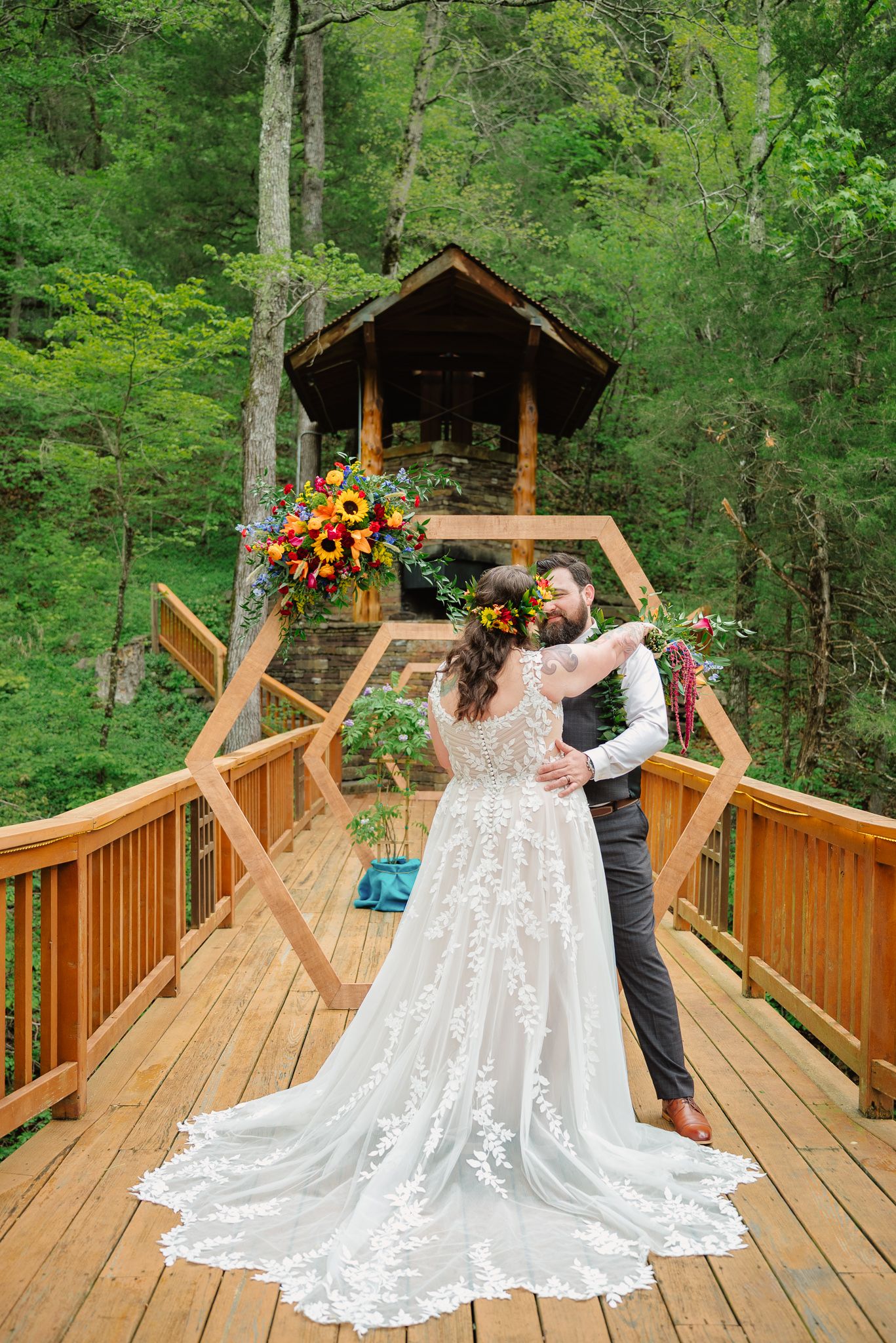 Bride and groom embrace on a wooden deck surrounded by lush forest with a treehouse and hexagonal arch backdrop.