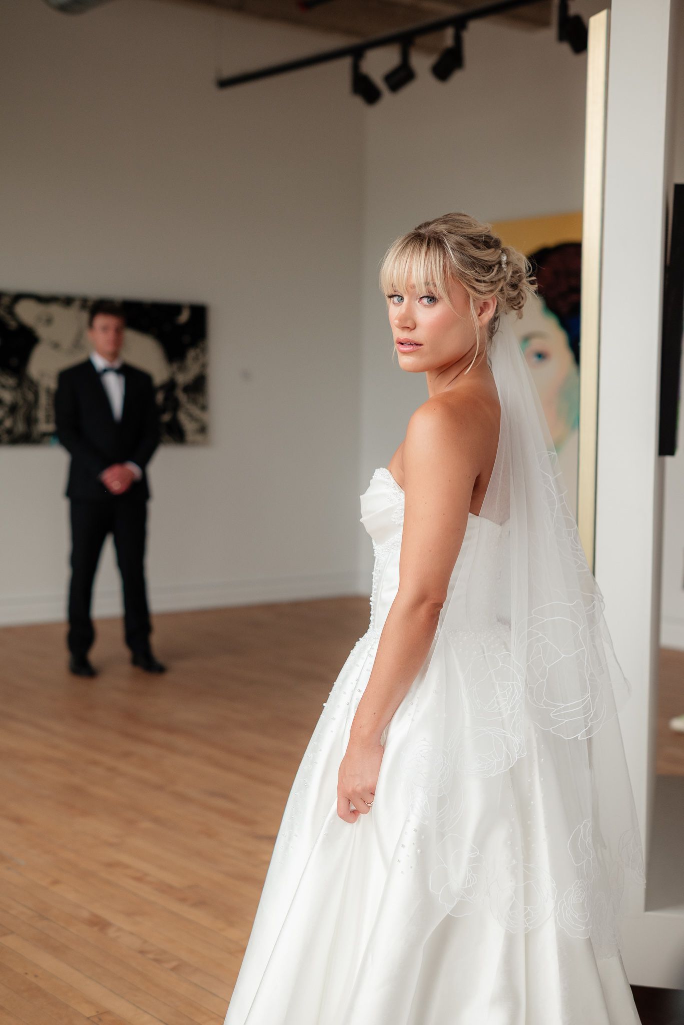 Bride in white strapless gown and veil stands elegantly indoors as groom in tuxedo stands in background.