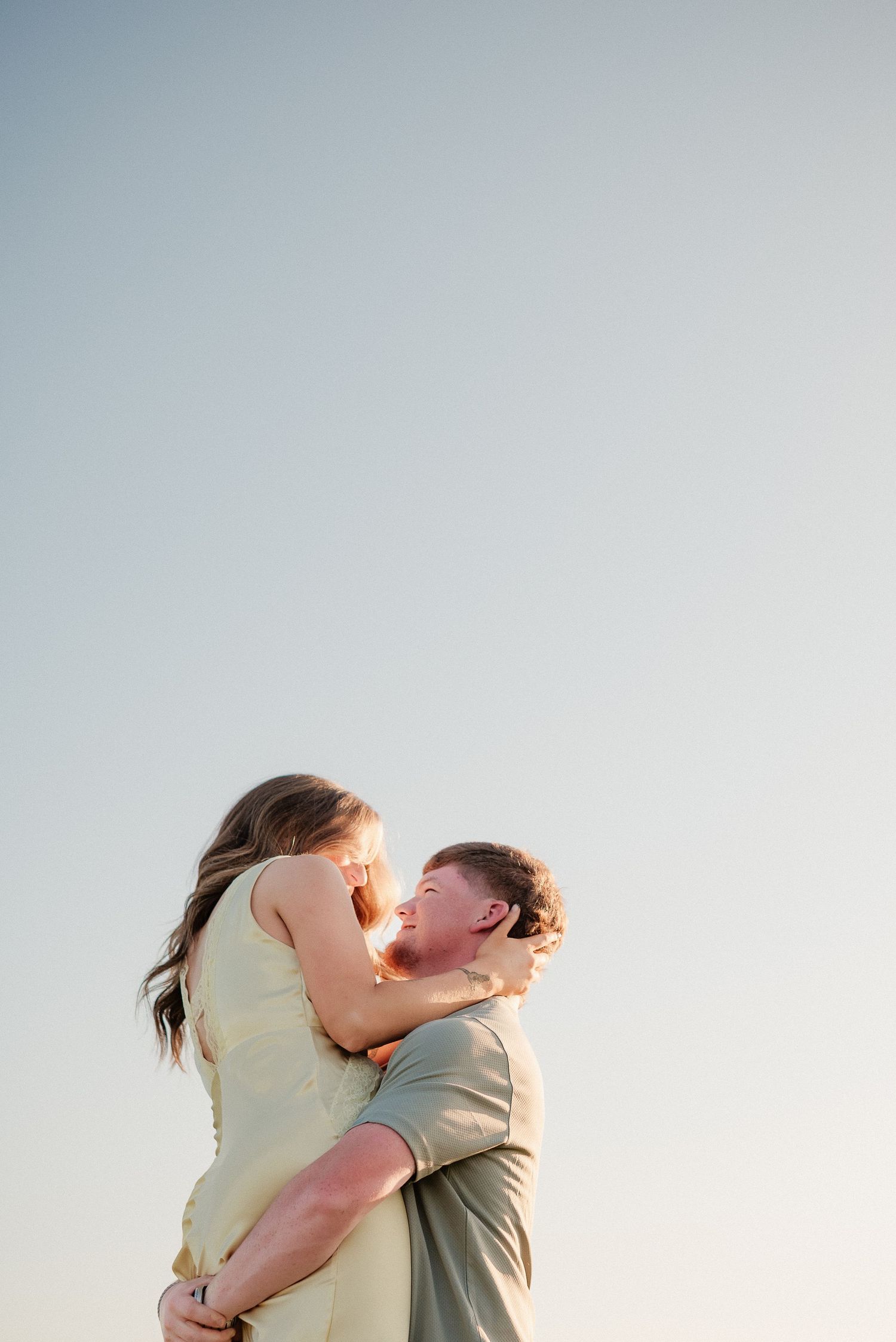 A joyful couple embraces outdoors, the man lifting the woman as they laugh together against a clear blue sky.
