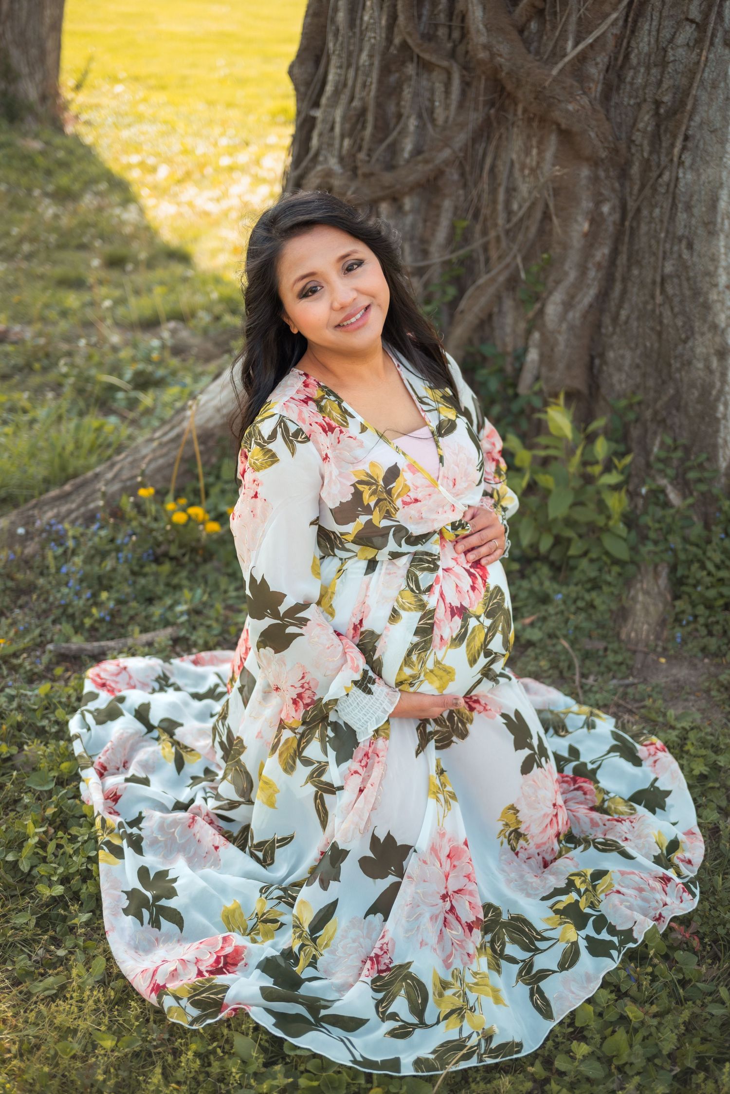Pregnant woman in floral maxi dress posing outdoors near tree, smiling warmly in a lush green garden setting.