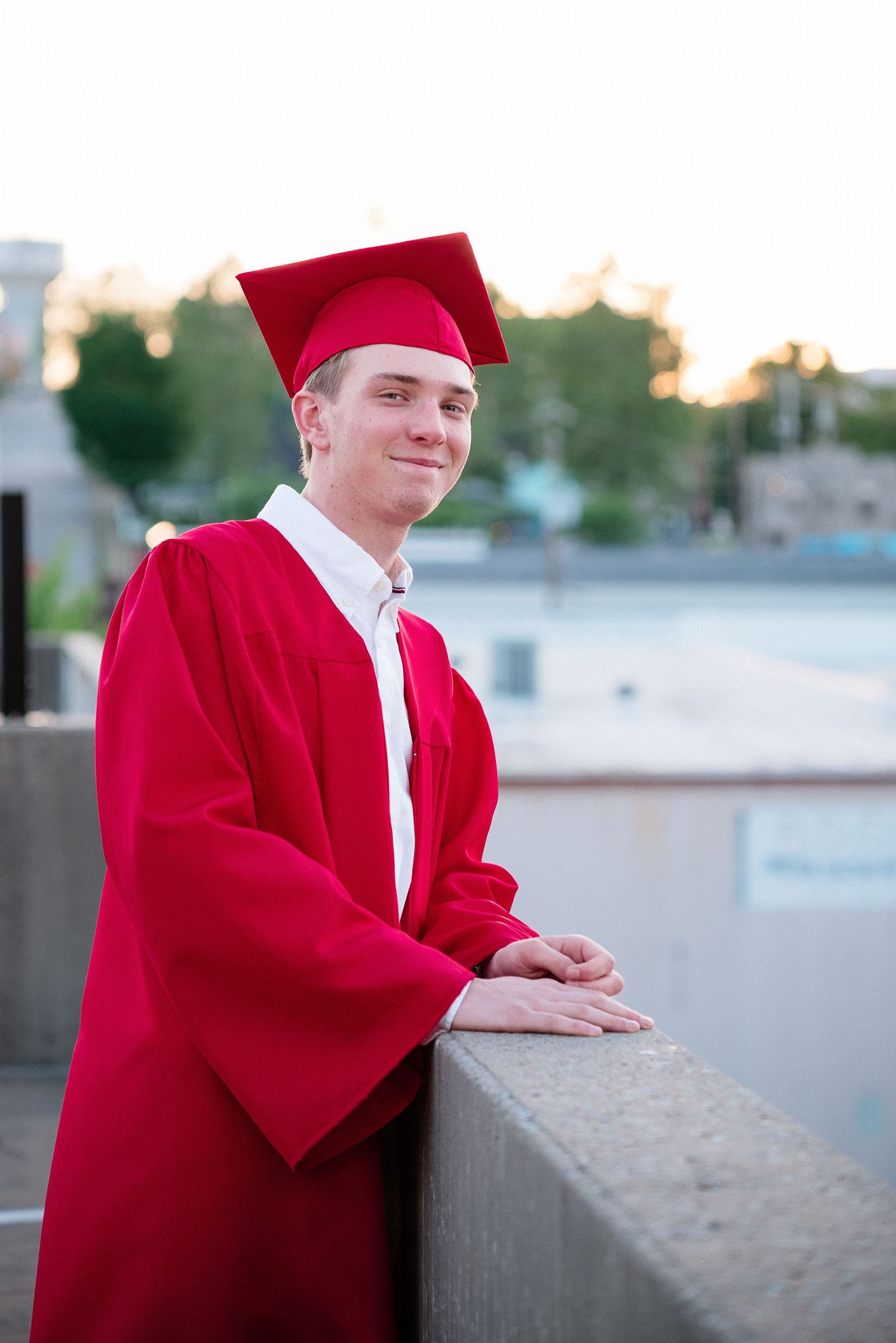Young man in red graduation cap and gown smiling while leaning on a concrete railing outdoors.