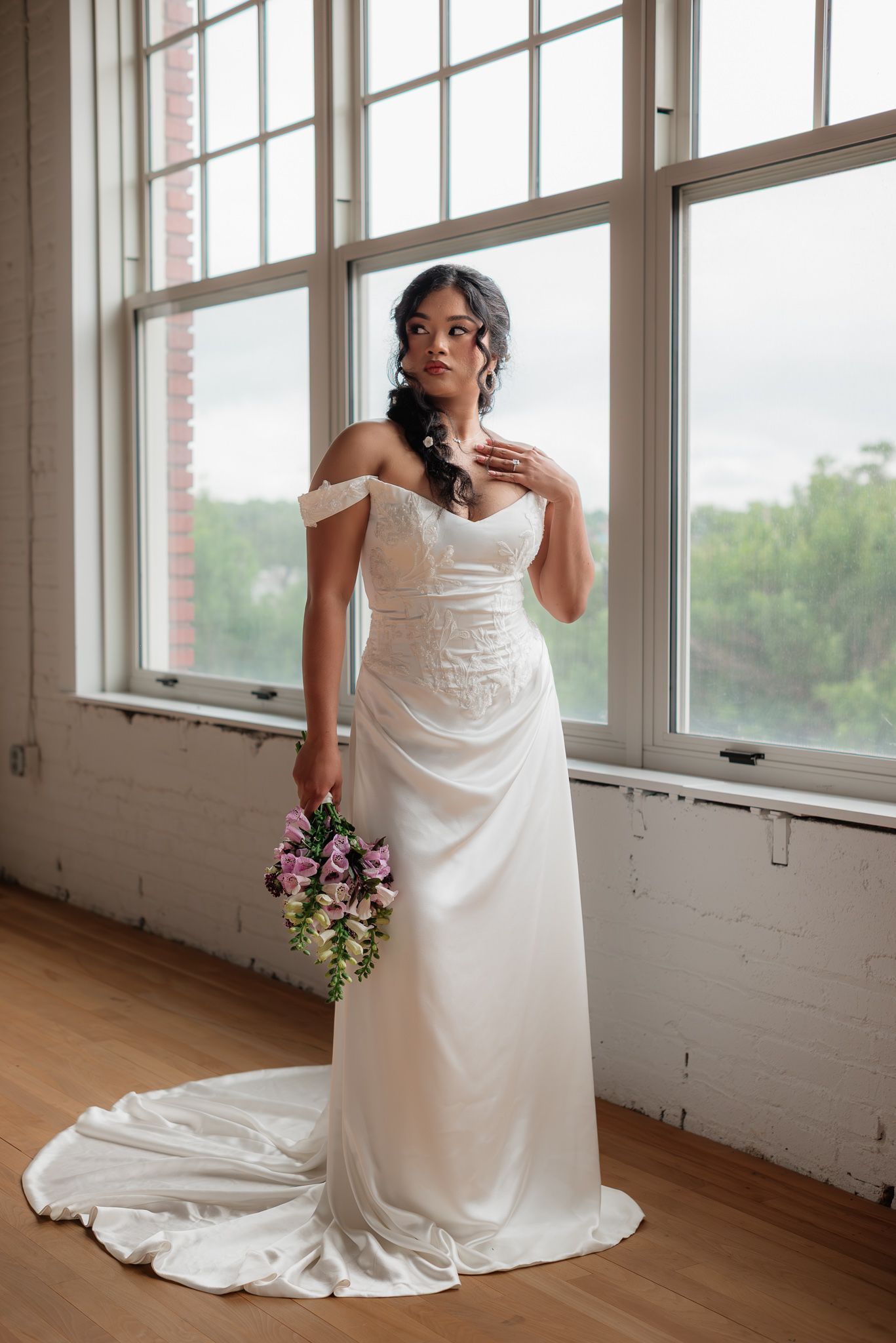 Bride in off-shoulder white satin gown with lace bodice holding pink bouquet, standing by large windows in brick venue.