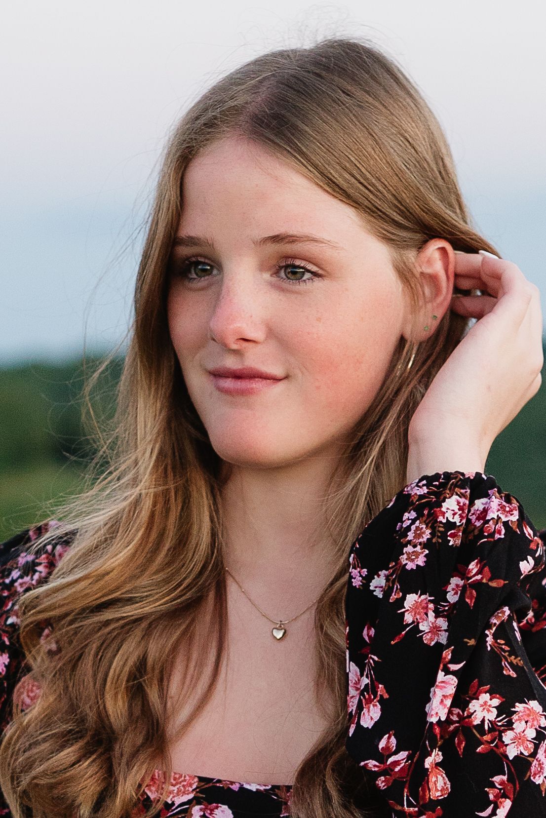 Young woman with long blonde hair wearing a black floral dress and heart necklace poses outdoors.