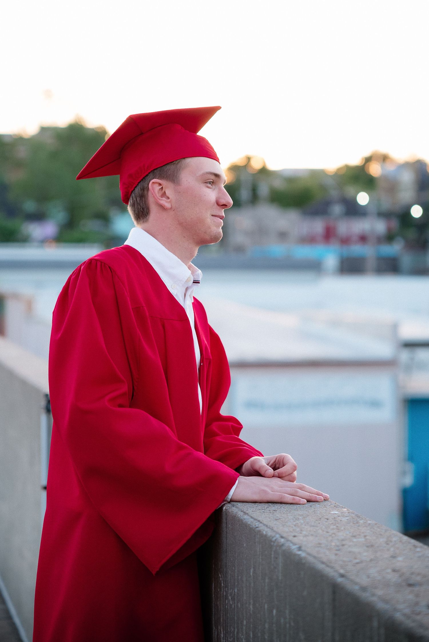 Young man in red graduation cap and gown leaning on a railing, looking into the distance outdoors.