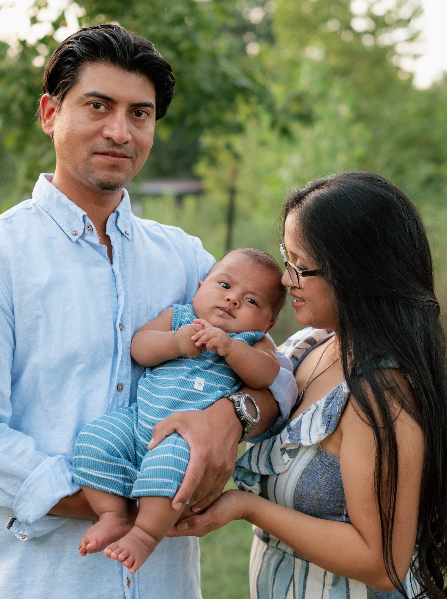 Young couple outdoors holding their newborn baby, smiling warmly in a lush green park setting.