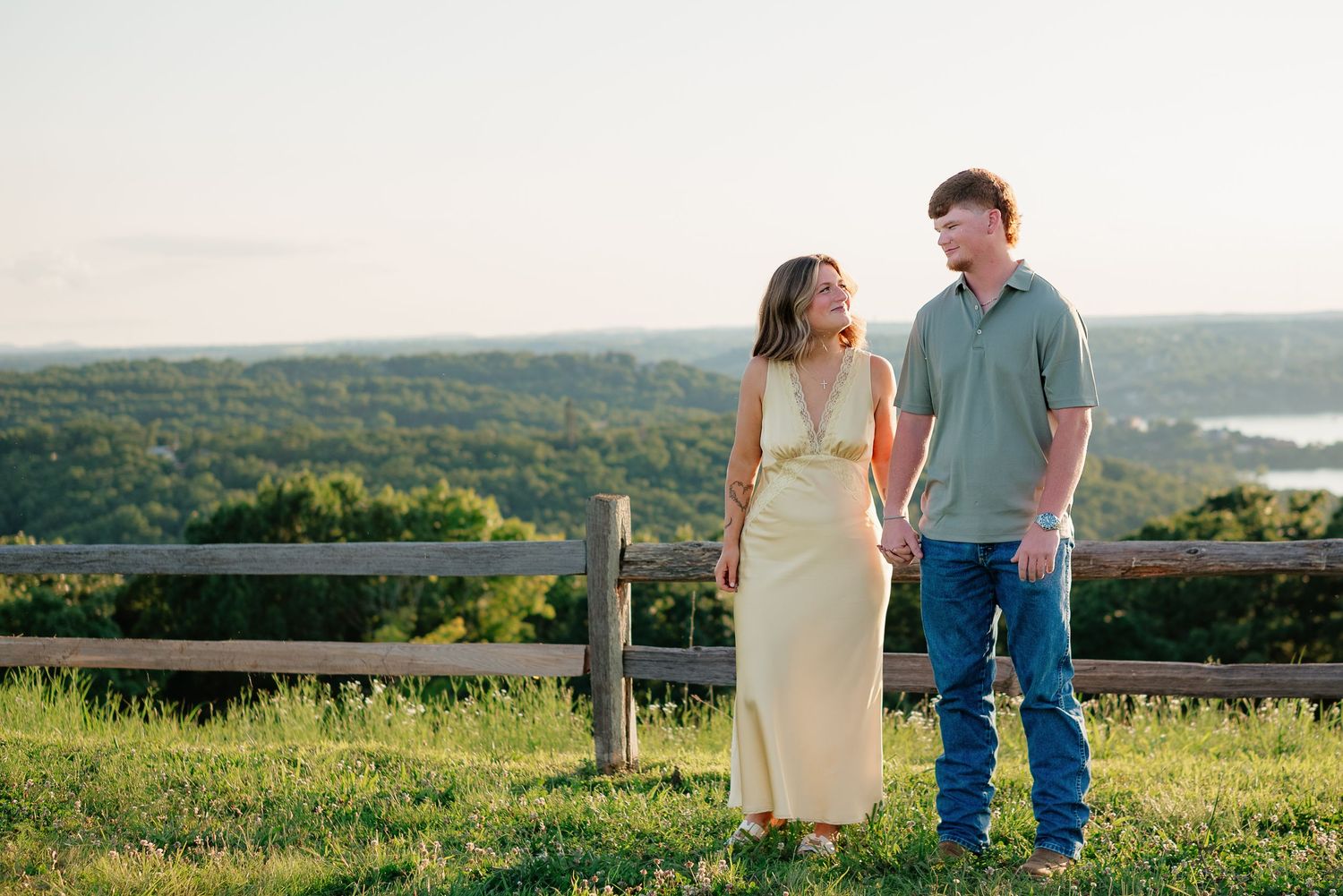 Couple holding hands in a yellow dress and blue shirt on a green hill with scenic lake views at sunset.