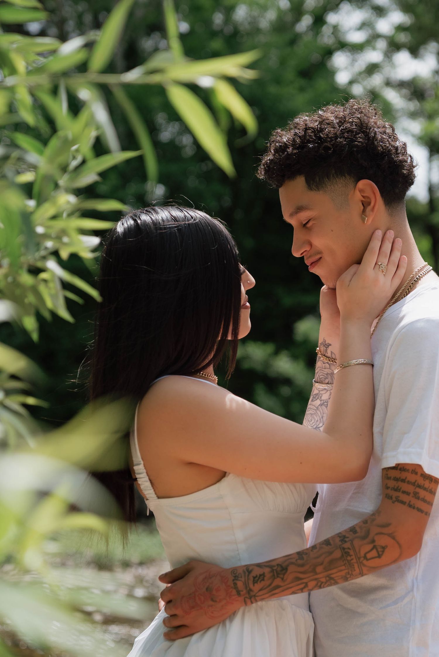 A tattooed couple embraces and smiles at each other outdoors, surrounded by lush green tropical foliage.