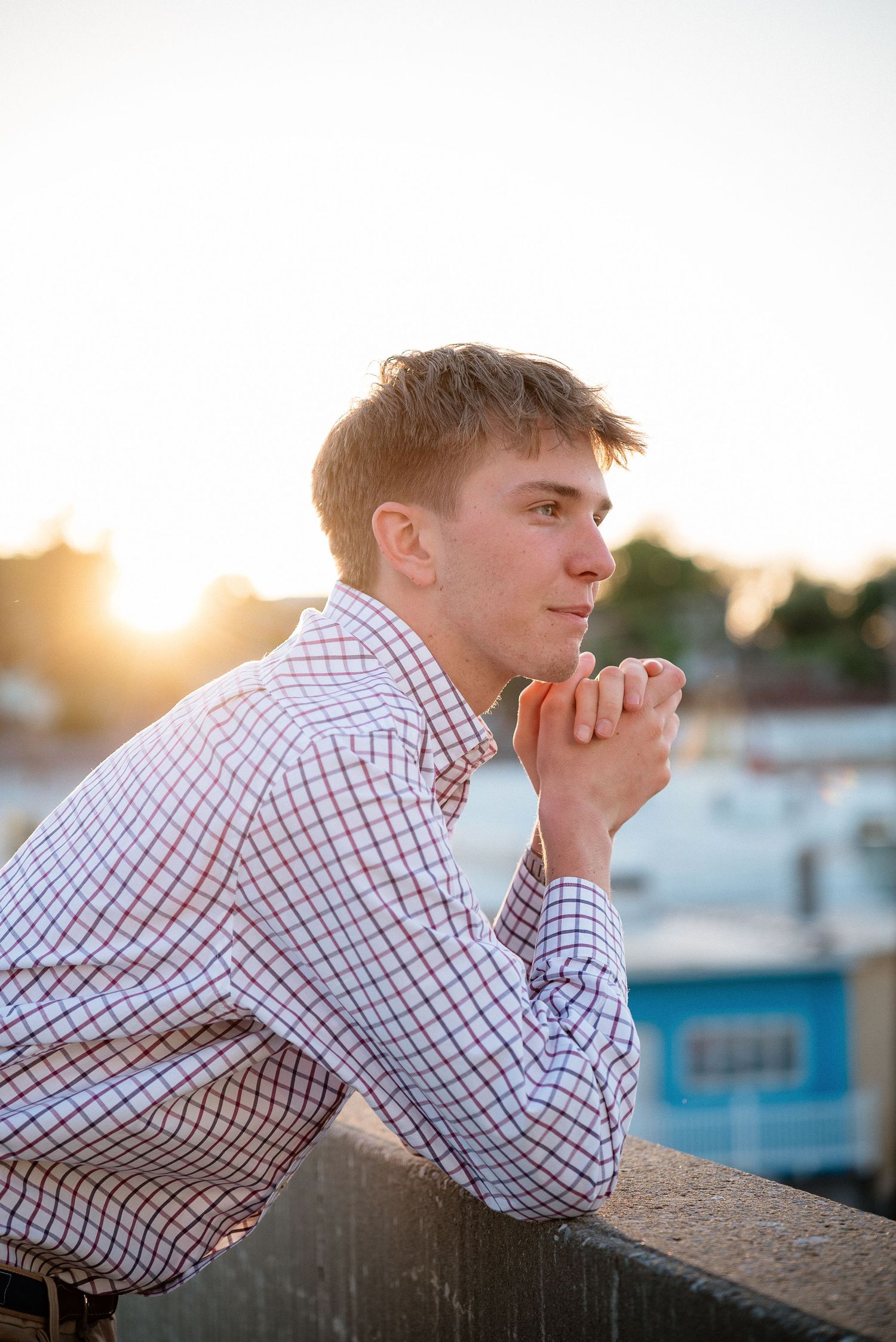 Young man in plaid shirt leaning on railing at sunset, hands clasped, gazing thoughtfully into the distance.