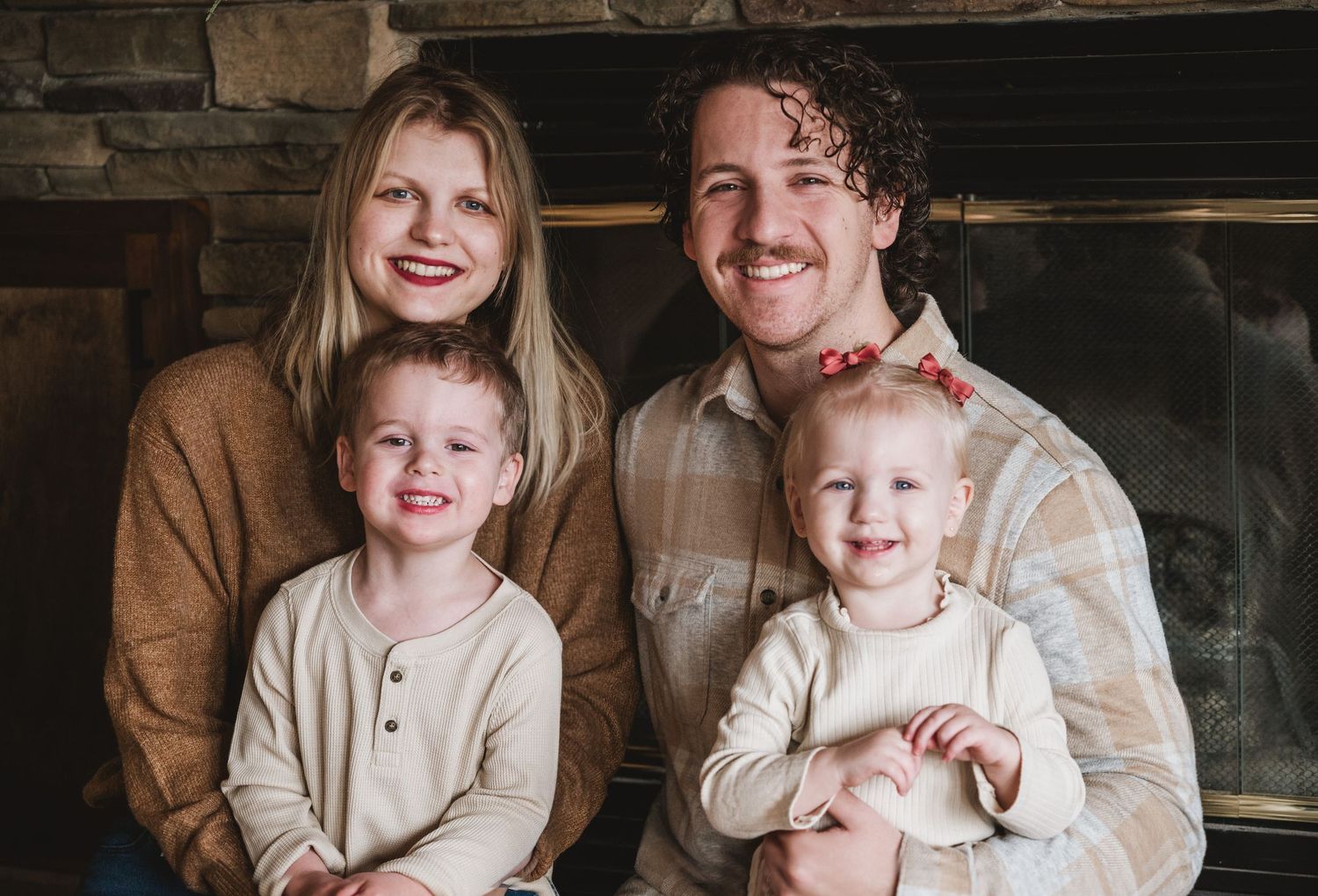 A smiling family of four poses warmly by a stone fireplace, featuring two young children in cream outfits.
