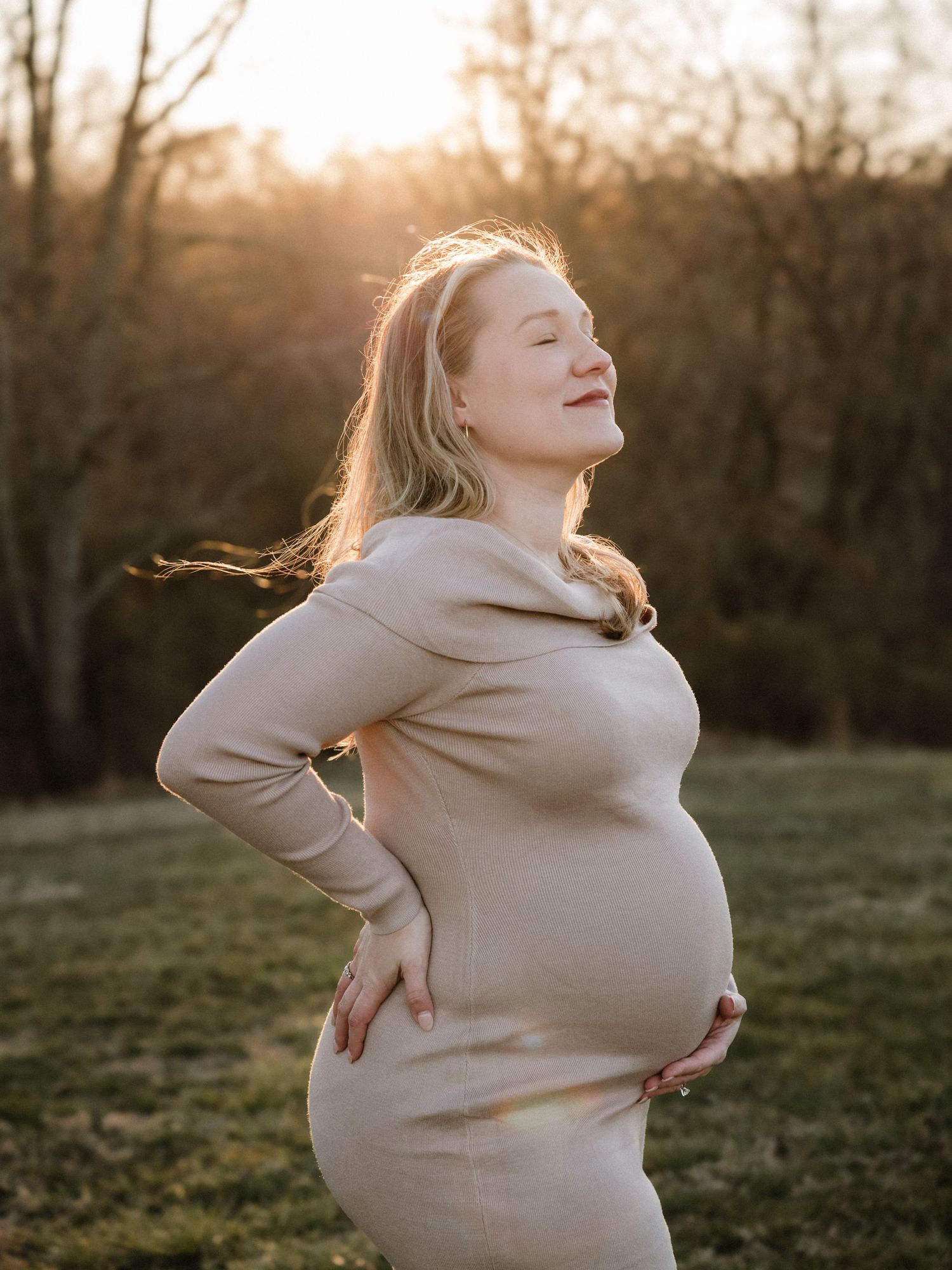 Pregnant woman in beige dress holding baby bump outdoors at golden hour sunset with trees in background.