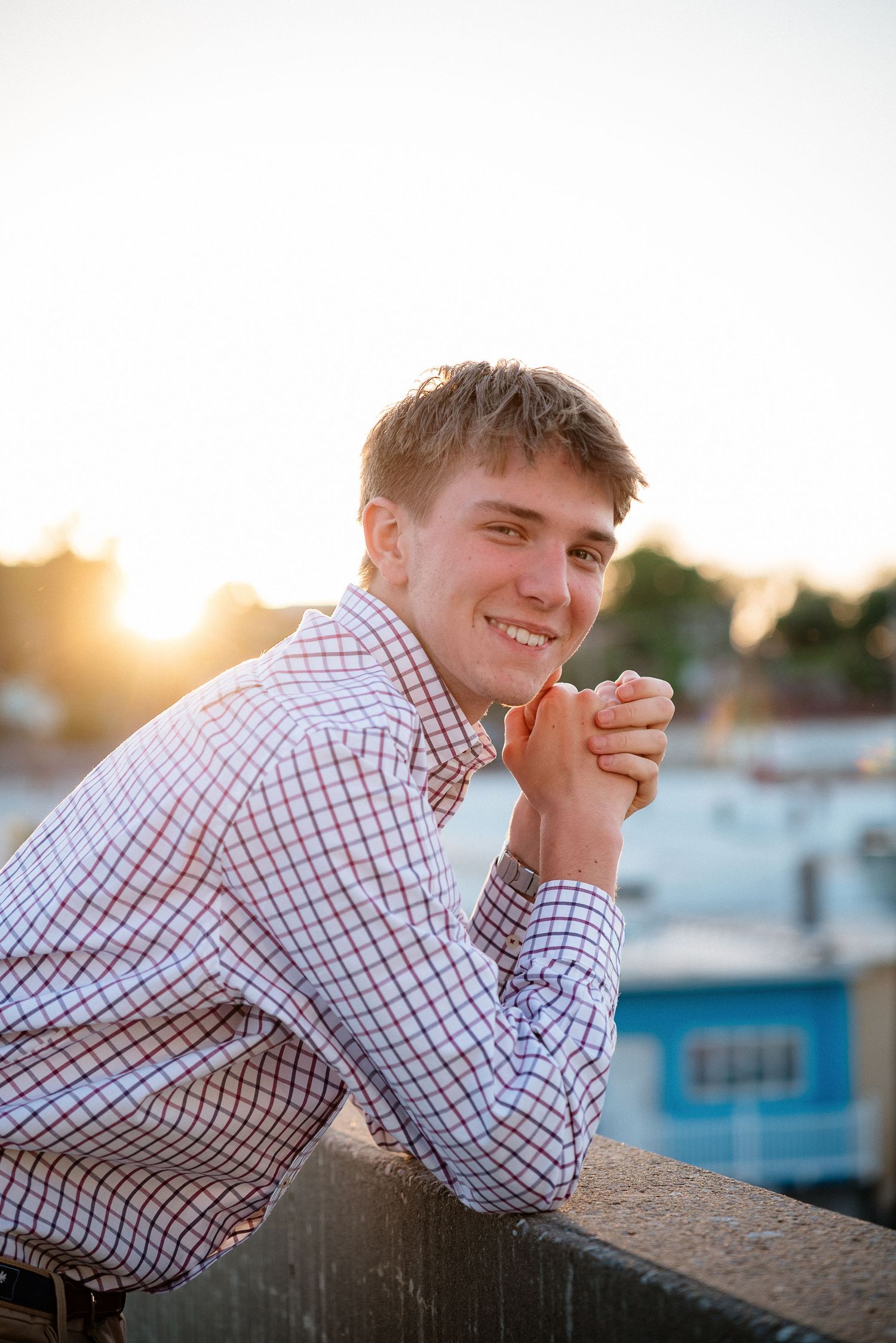 Smiling young man in plaid shirt leaning on railing at golden hour with cityscape background.