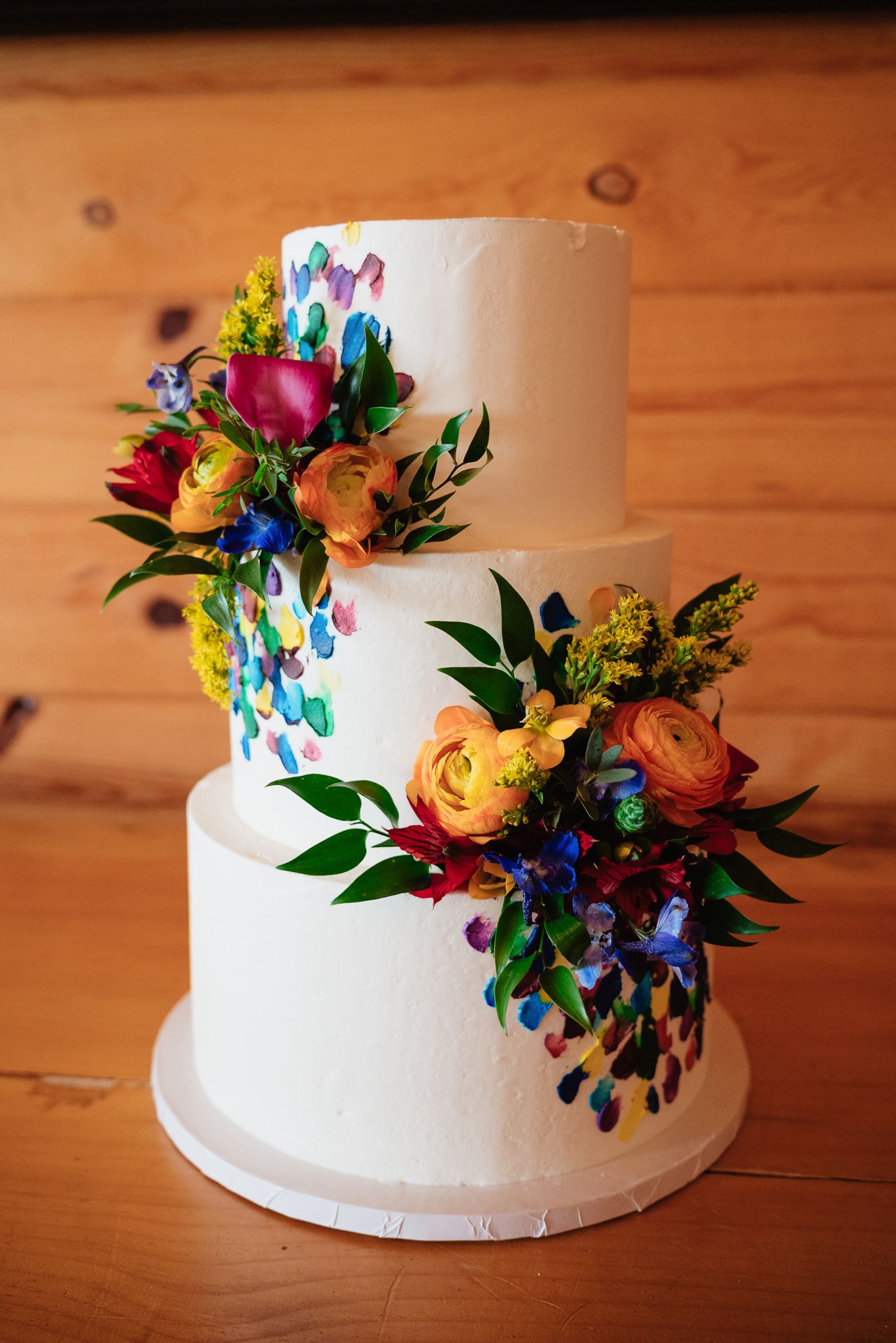 Three-tier white wedding cake adorned with colorful fresh flowers and painted butterfly details on wooden background.