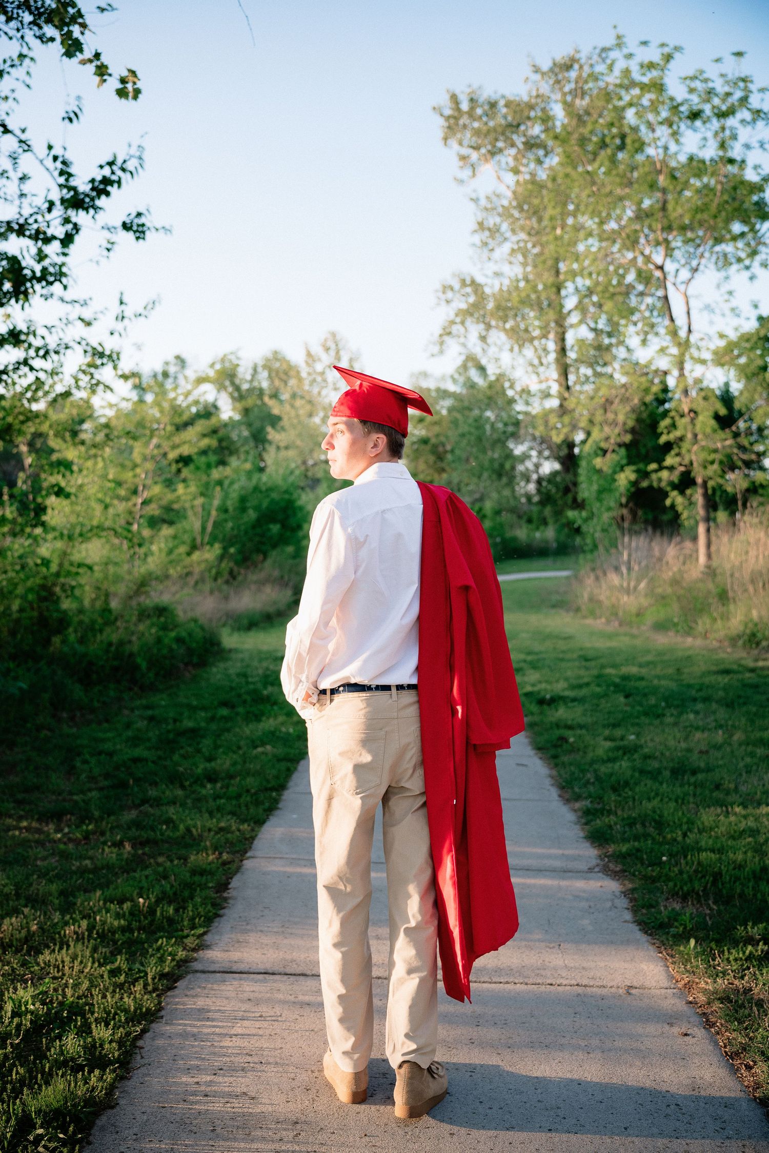 Graduate in red cap and gown stands on a tree-lined path, gazing forward in a reflective outdoor portrait.