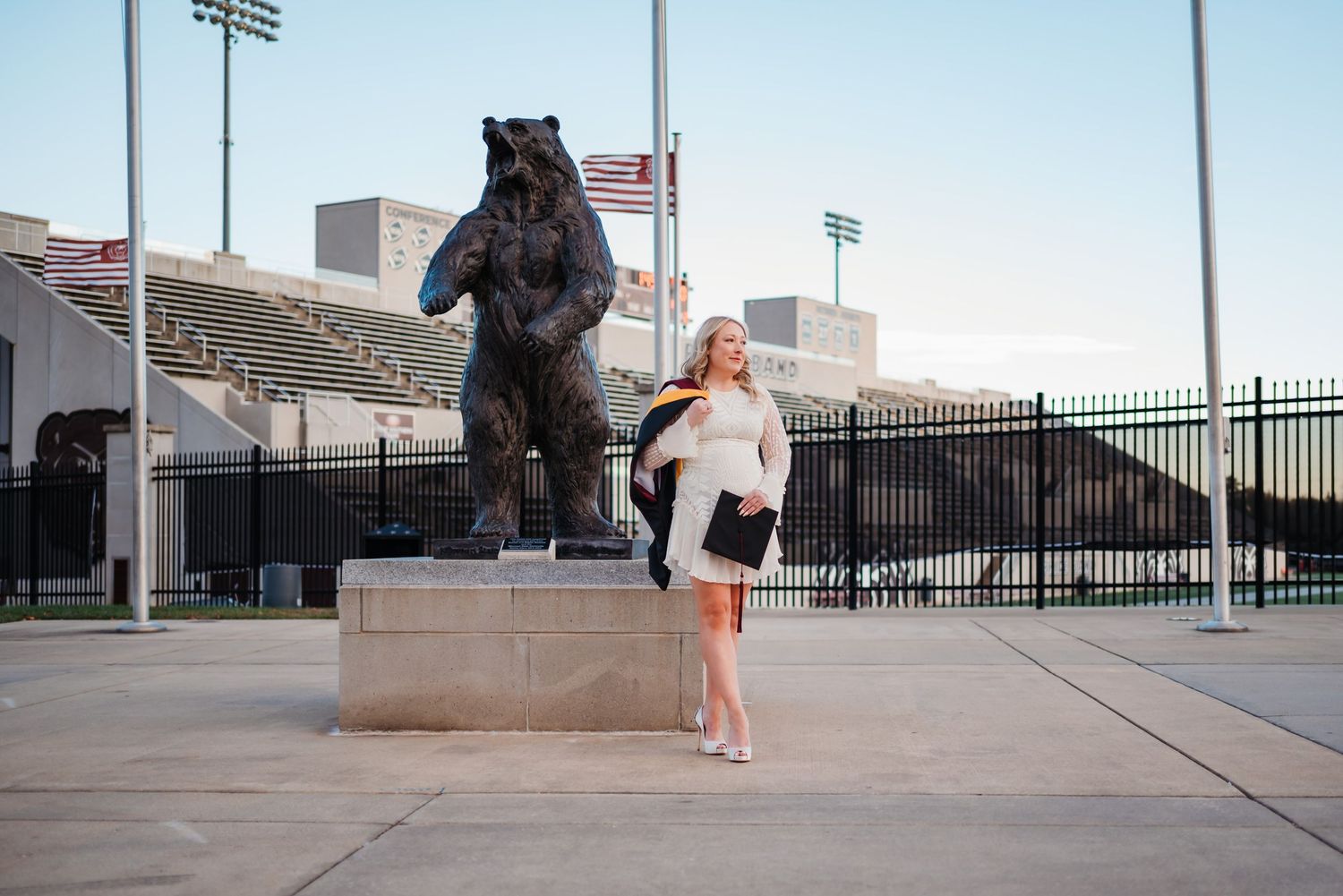 Blonde woman posing next to a bear statue outside a football stadium with American flags in background.