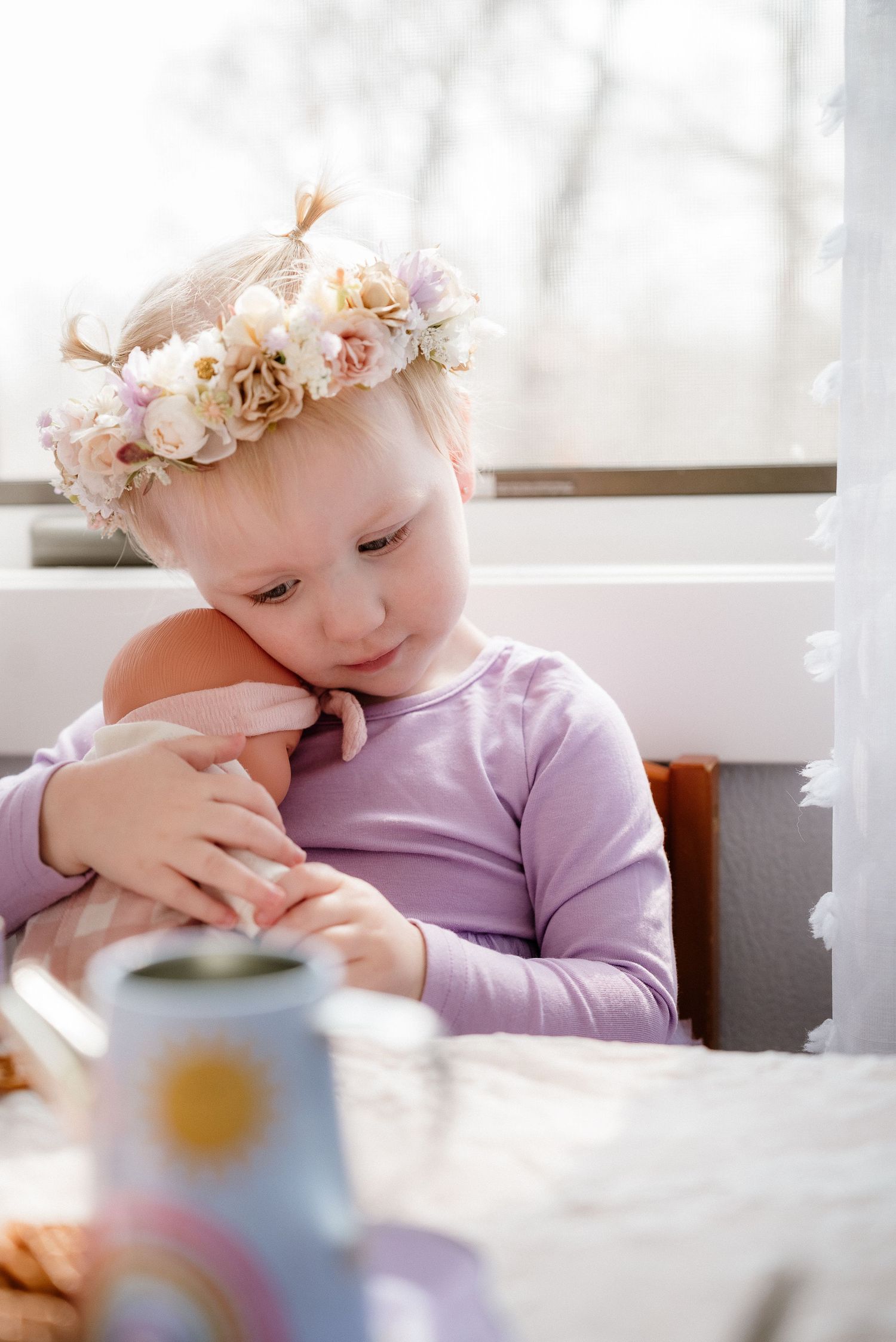Young girl wearing floral crown tenderly holds baby doll at sunny table, dressed in lavender top.