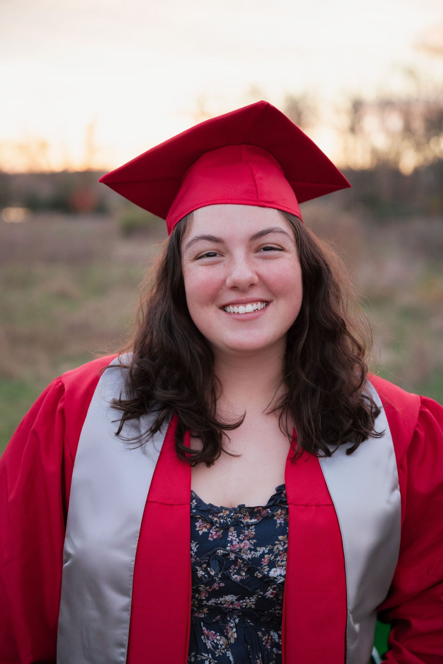 Smiling young woman in red graduation cap and gown posing outdoors at sunset.